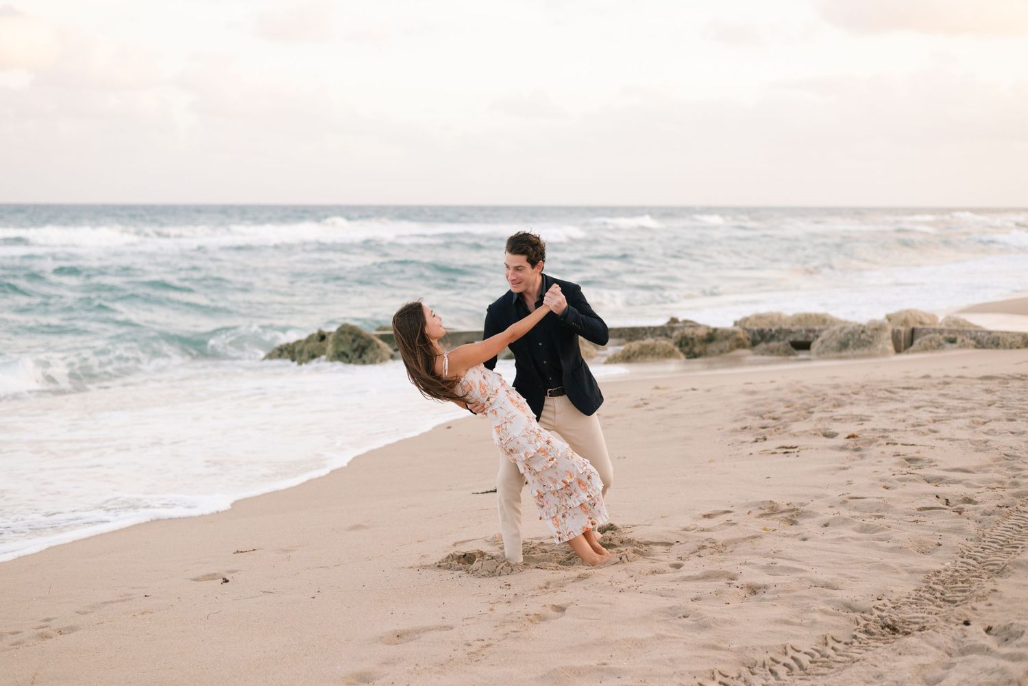 Couple playfully dancing together on a sandy beach at sunset during a romantic photoshoot.