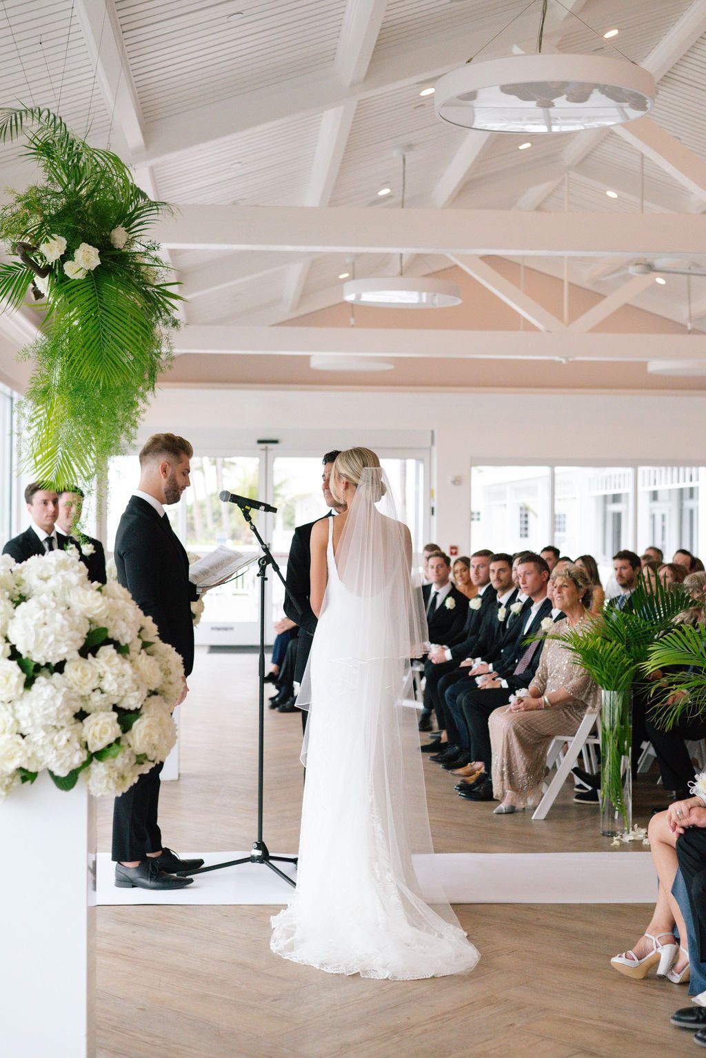 Elegant wedding ceremony in bright white room with vaulted ceiling and tropical palm decor.