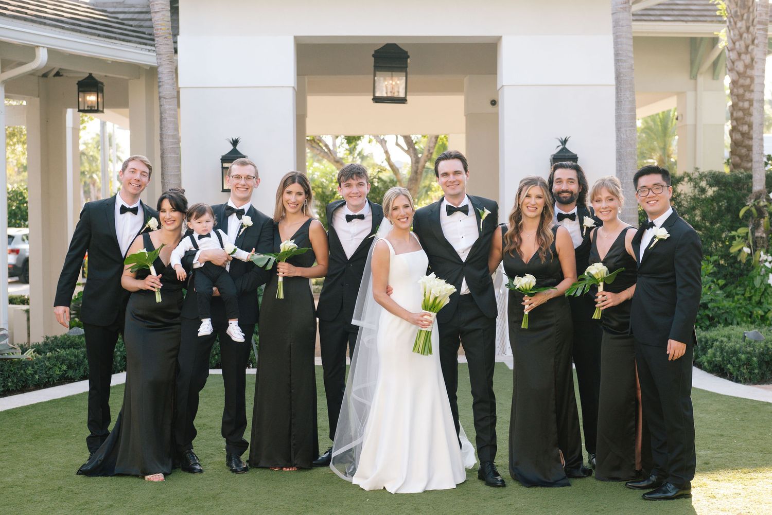 Wedding party in formal black attire and white dress gathered on lawn.