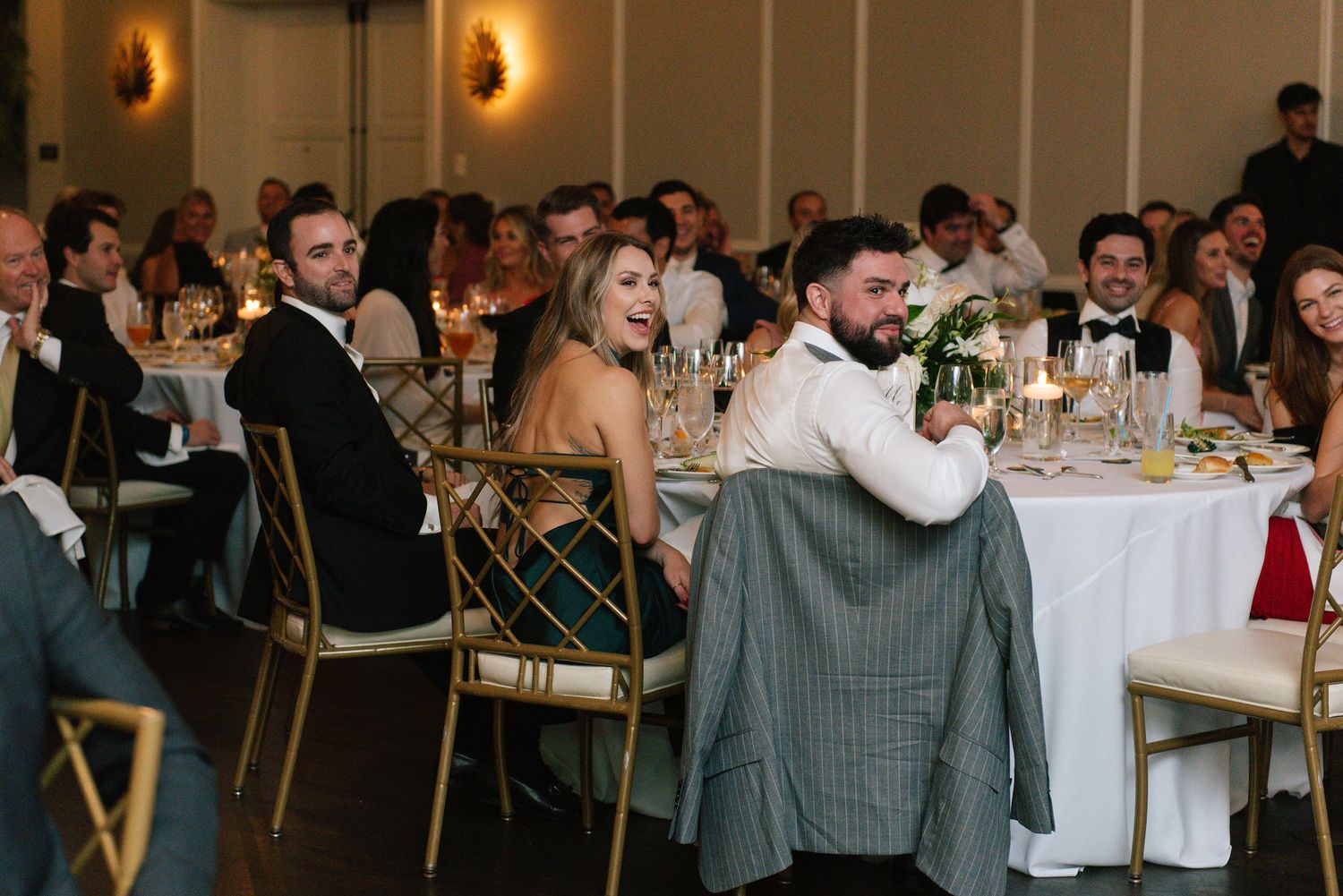 Wedding reception guests seated at round tables in an elegant ballroom with warm lighting.