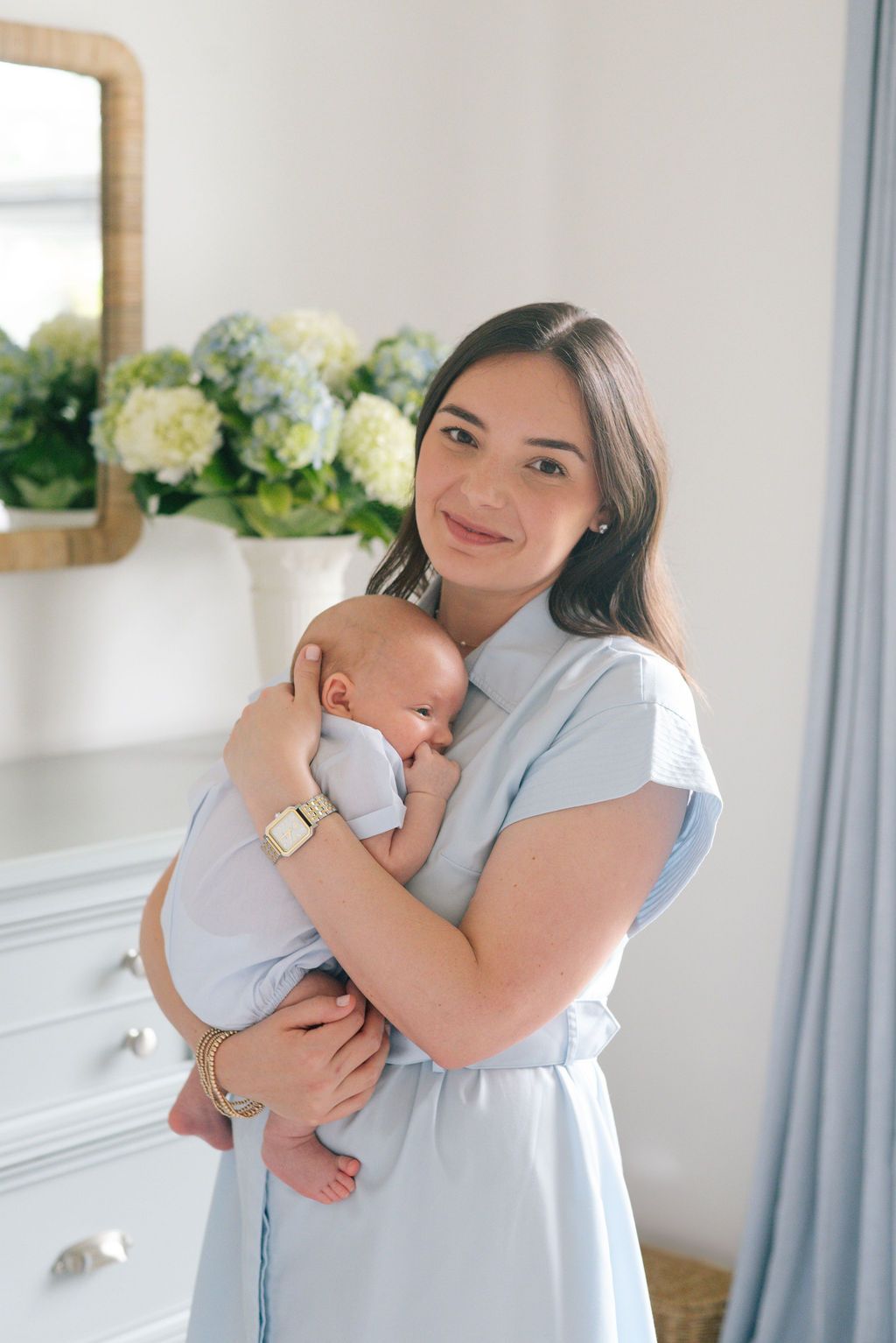 Mother in light blue dress holding sleeping newborn baby in front of hydrangea decorations.