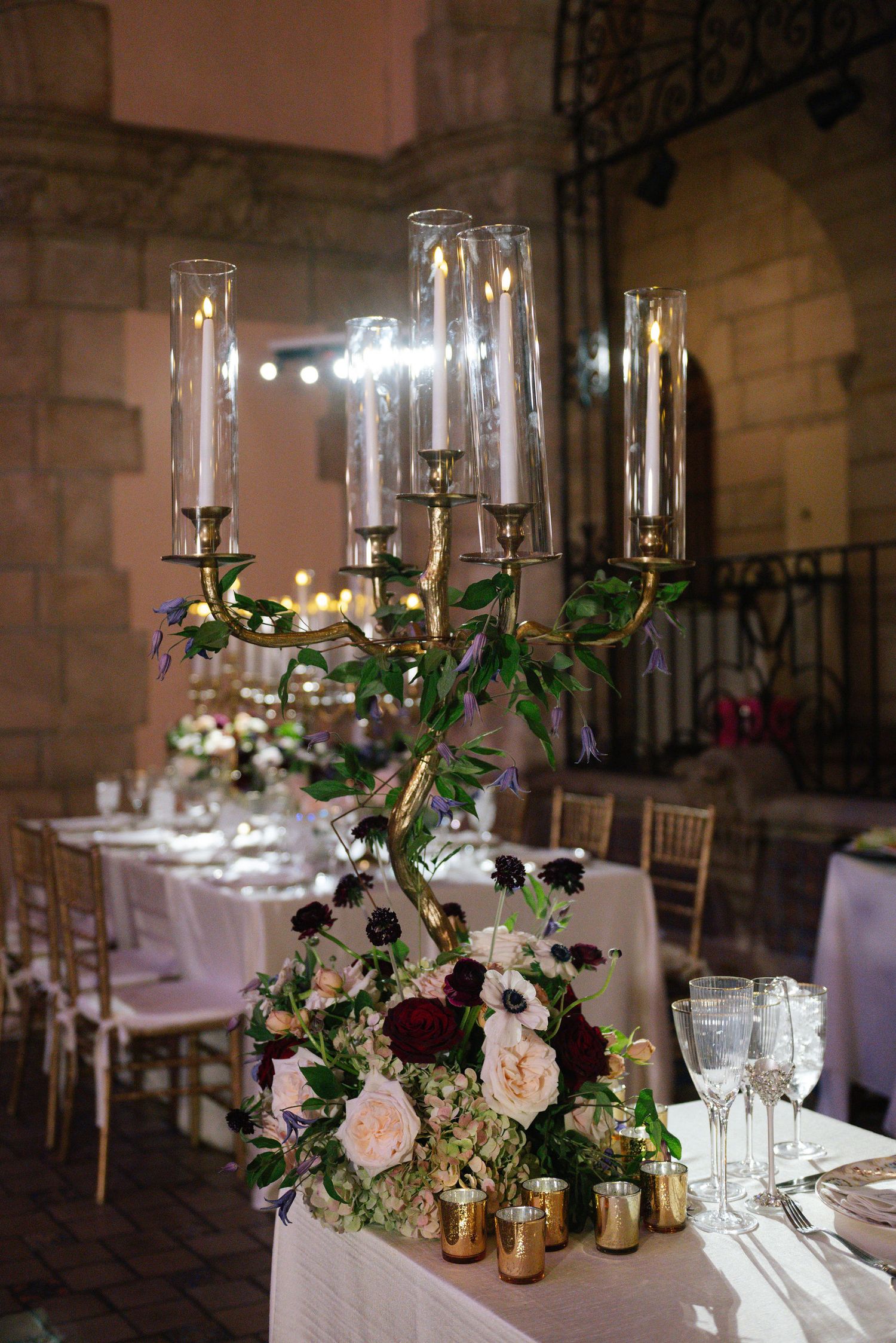 Elegant wedding table setting with tall glass candleholders and romantic floral arrangements in white and burgundy.