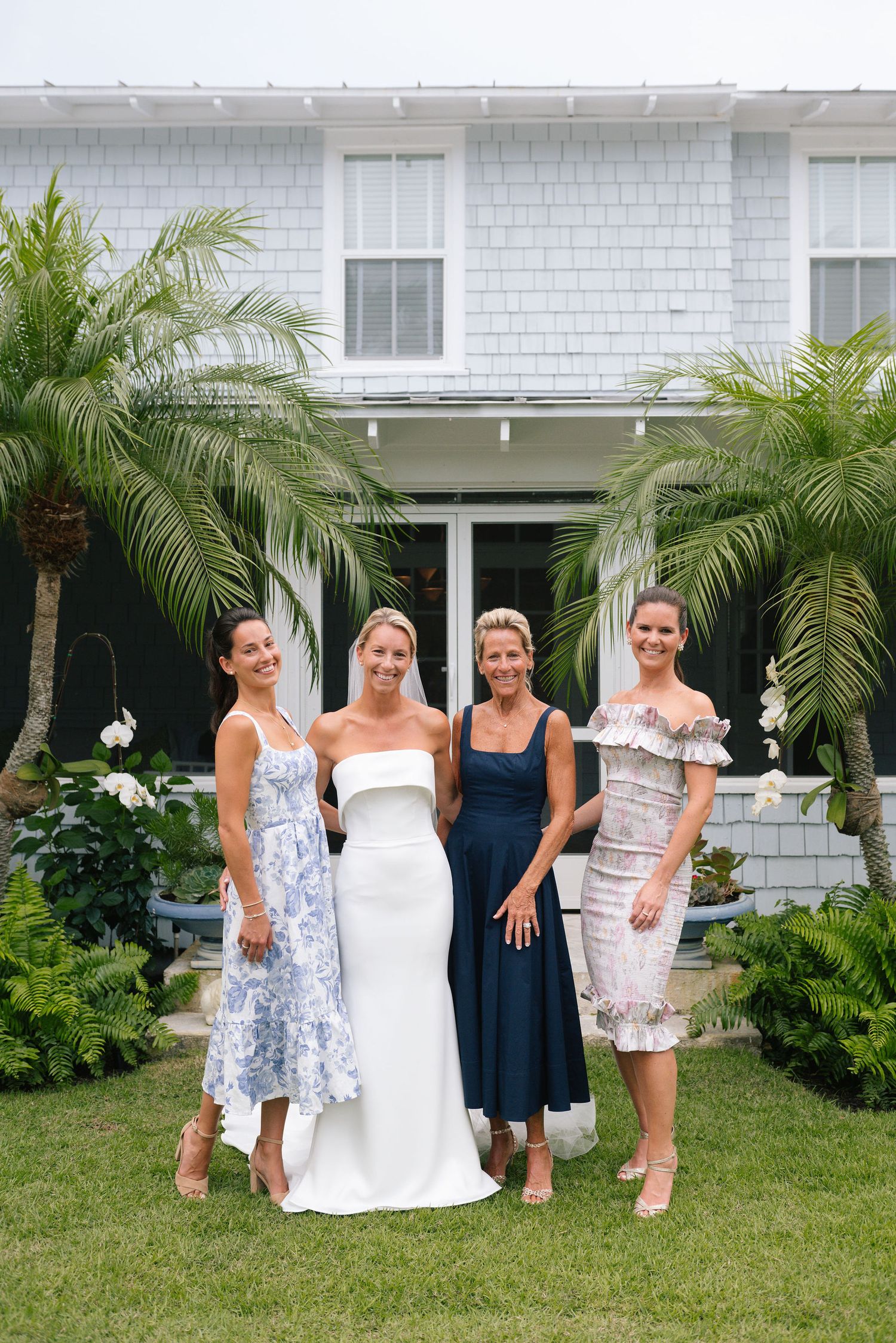 Wedding party in blue and white dresses poses on lawn in front of coastal-style house with palm trees.