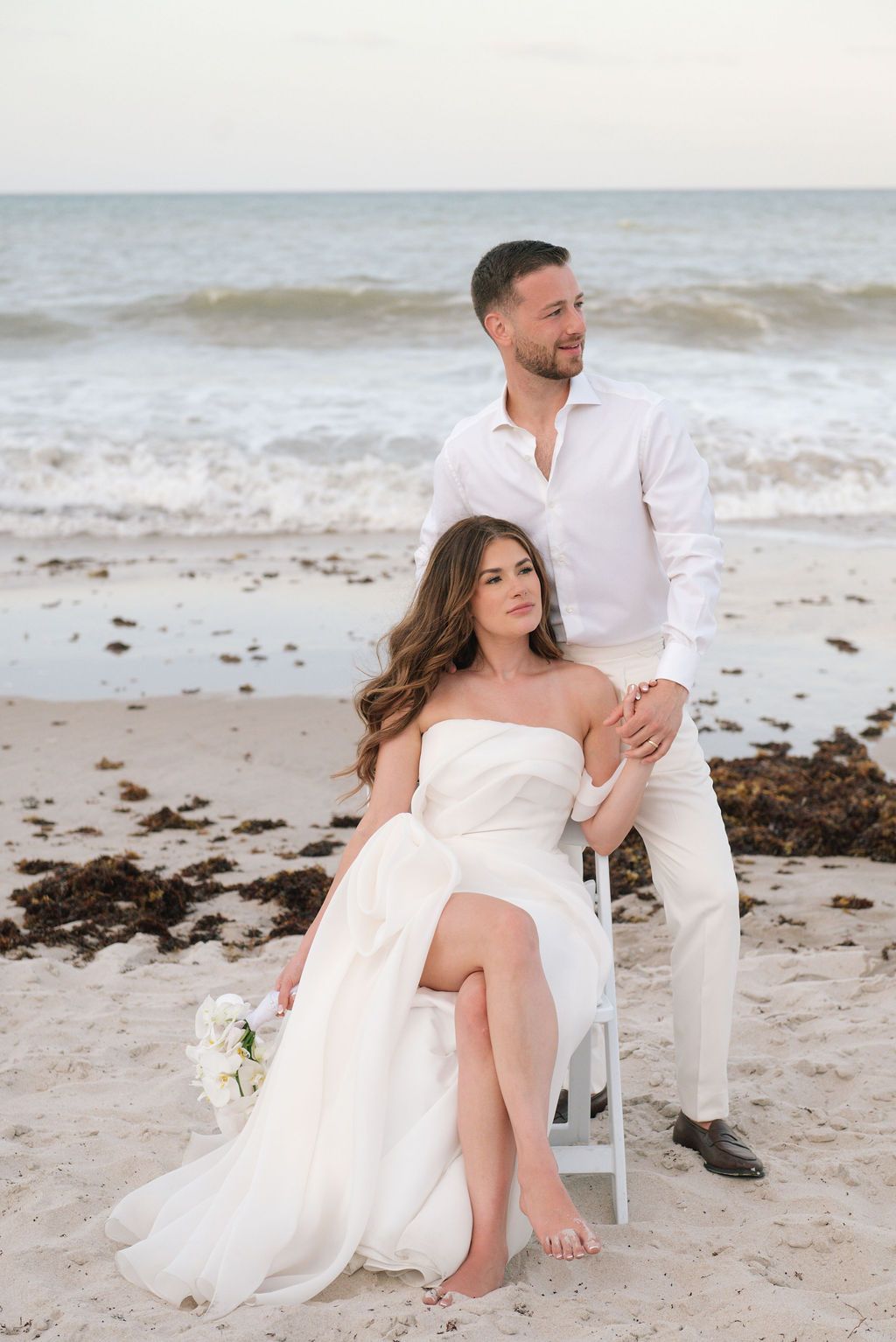 Artistic beach wedding portraits with couple seated on white chair by the shore.