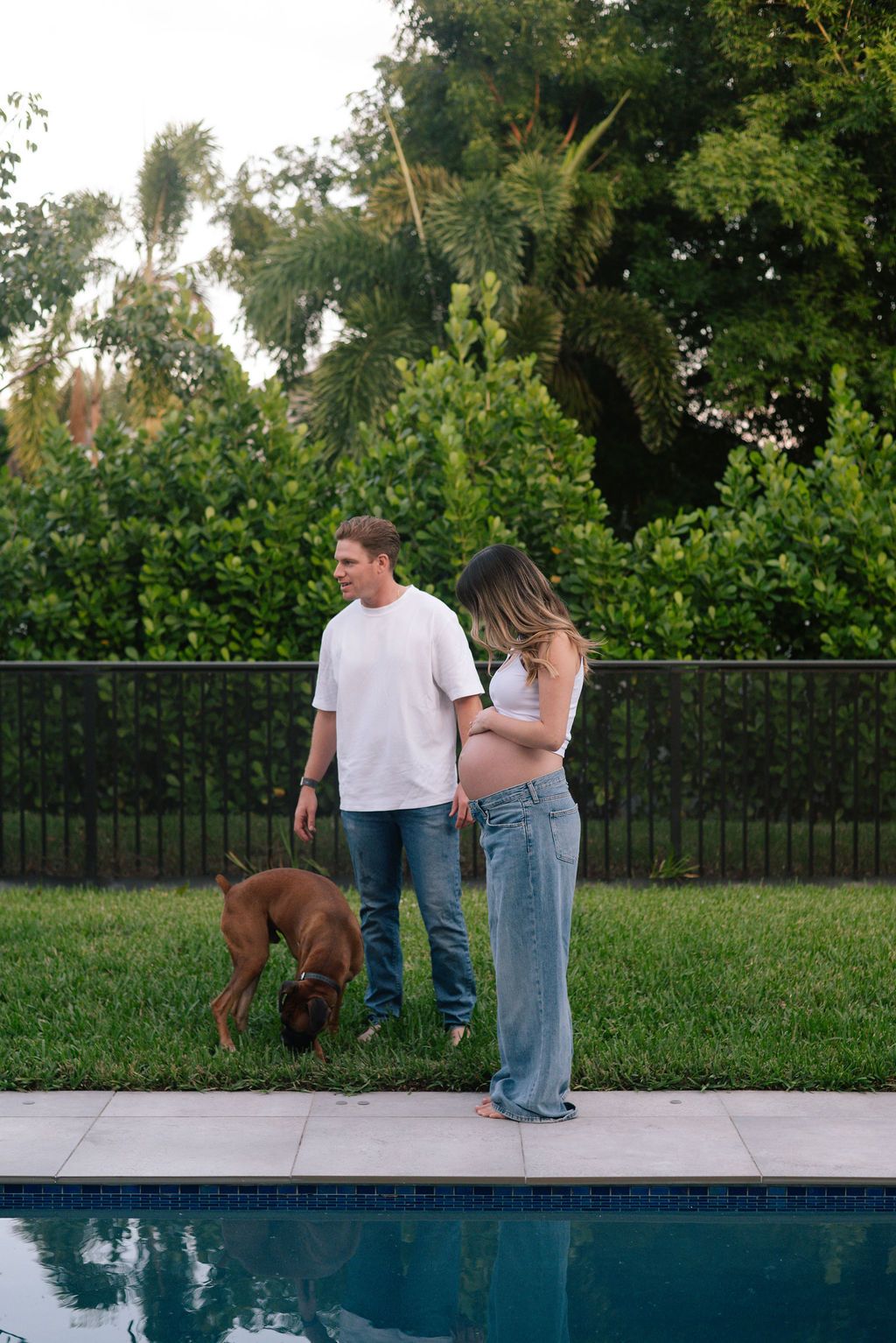 A couple and their dog stand near a swimming pool surrounded by lush greenery.