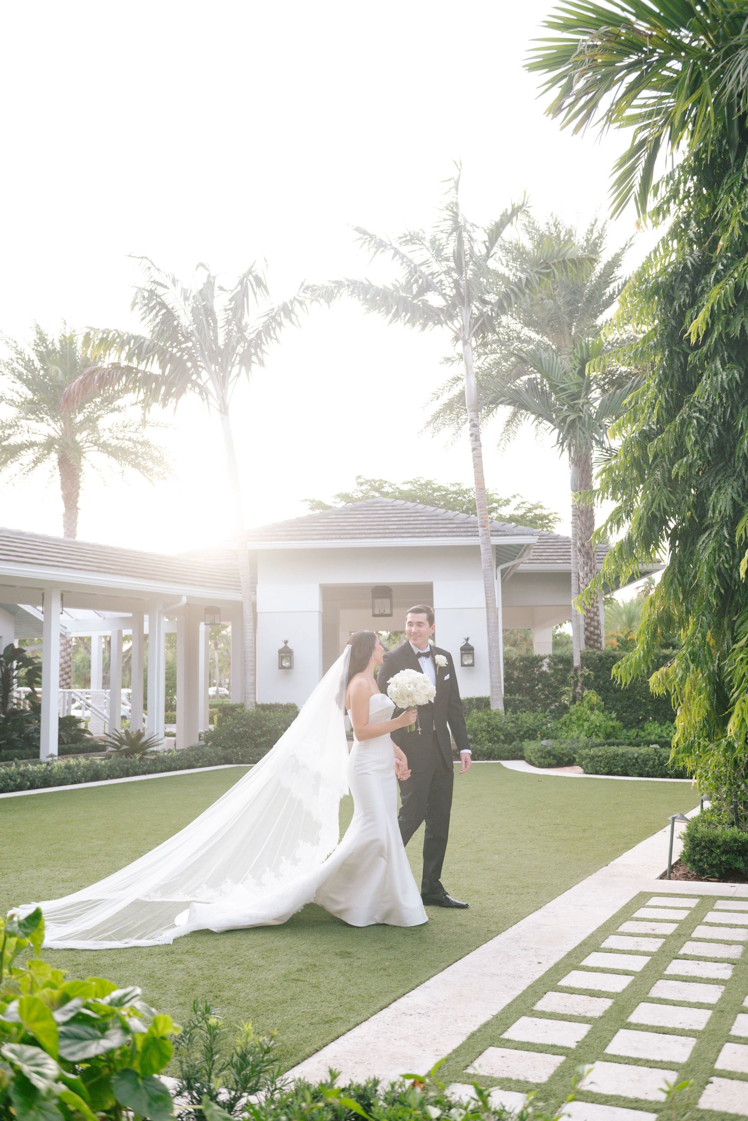 Wedding couple walks along a palm-lined pathway with flowing white dress trailing behind.