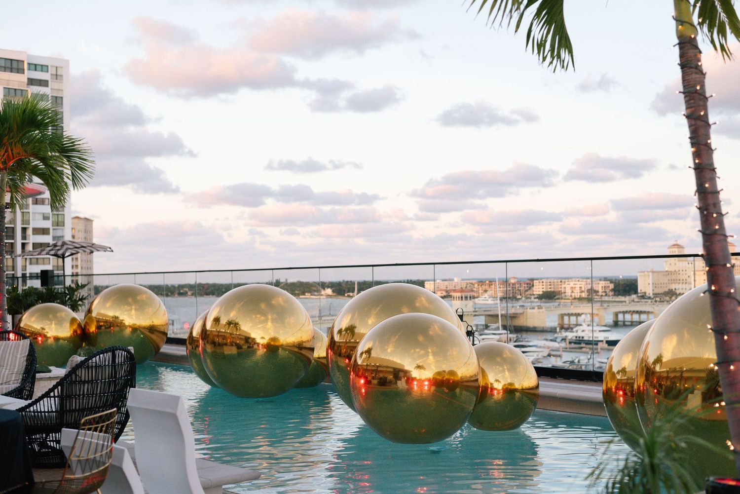 Large golden spherical decorations float in pool with palm trees and city skyline at sunset.