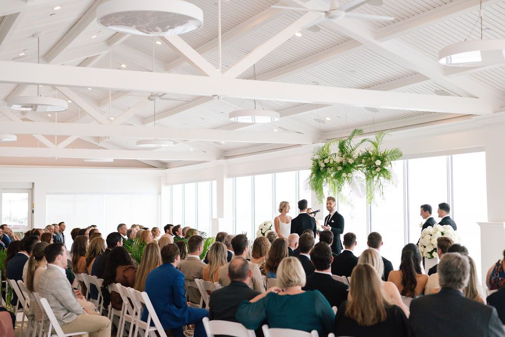 Wide view of wedding ceremony guests seated in bright white venue with exposed beam ceiling.