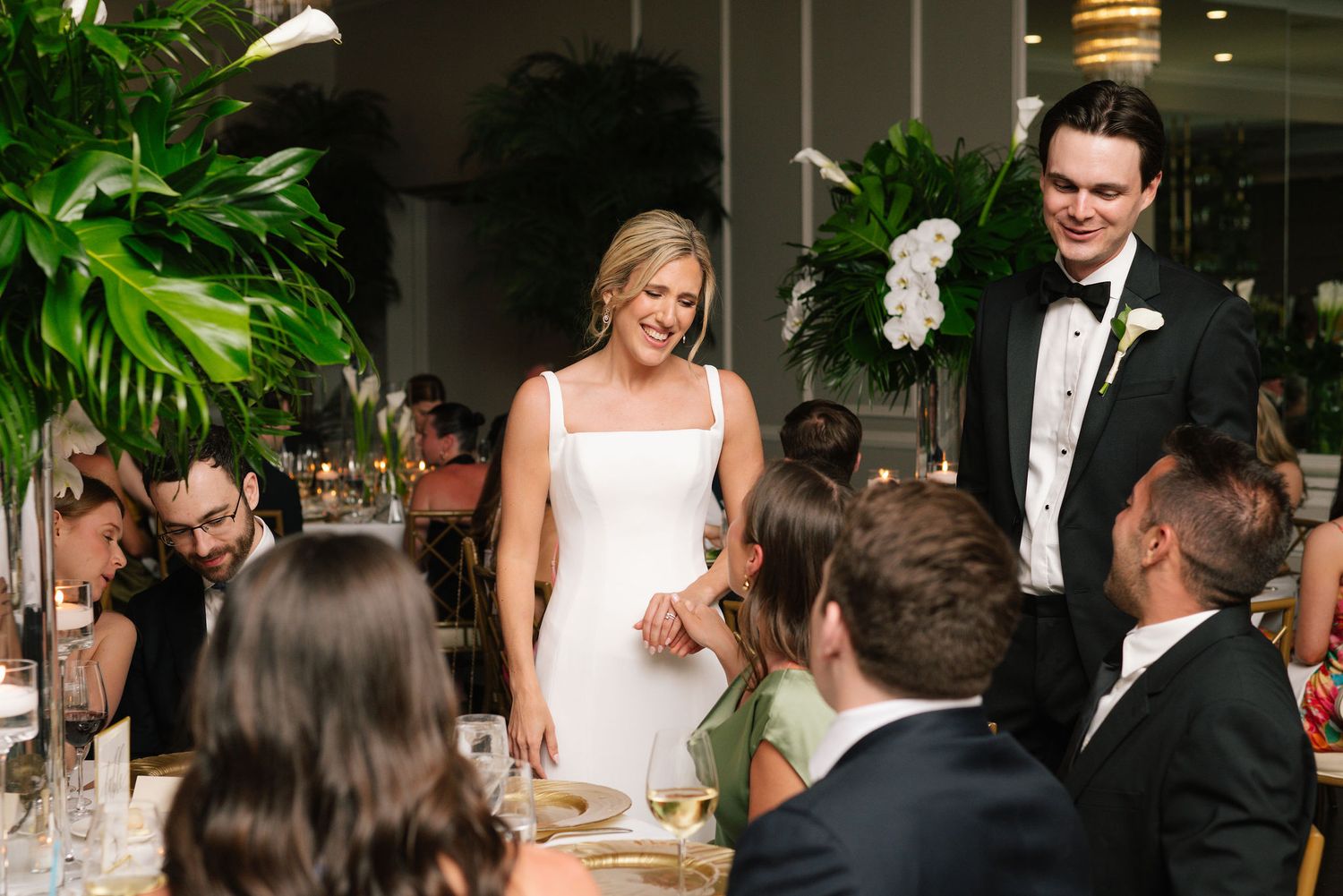 Elegant wedding reception setup with tropical greenery and warm lighting in an indoor venue.