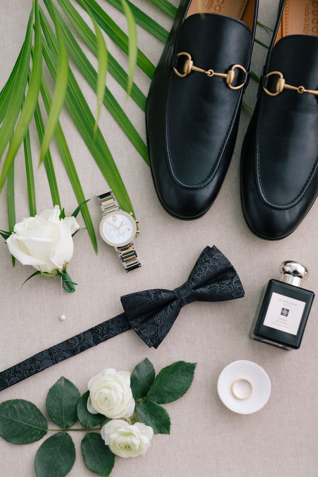 Flatlay of black leather shoes, white roses, and perfume bottles on neutral background.