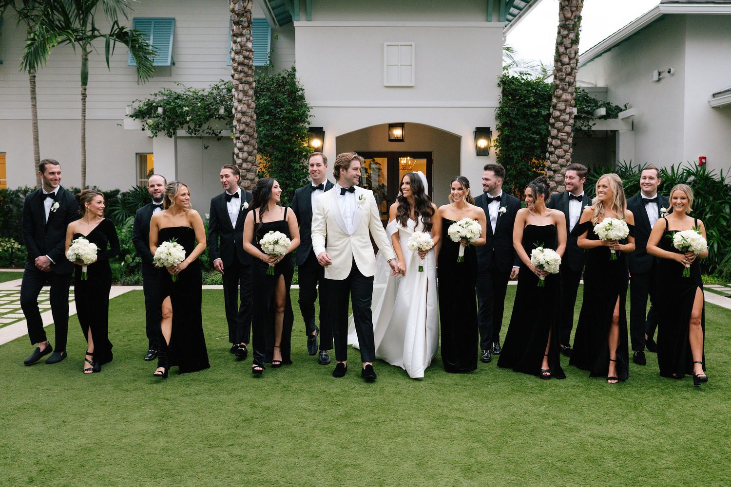 Wedding party members arranged for formal photos on grass with palm trees in background.