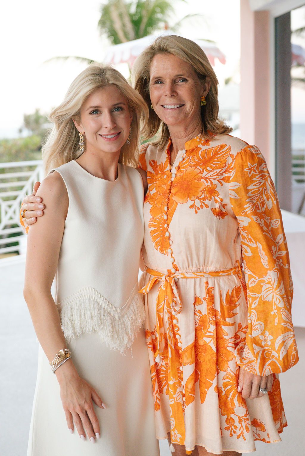 Two women in contrasting outfits of white and orange floral print on a coastal balcony.