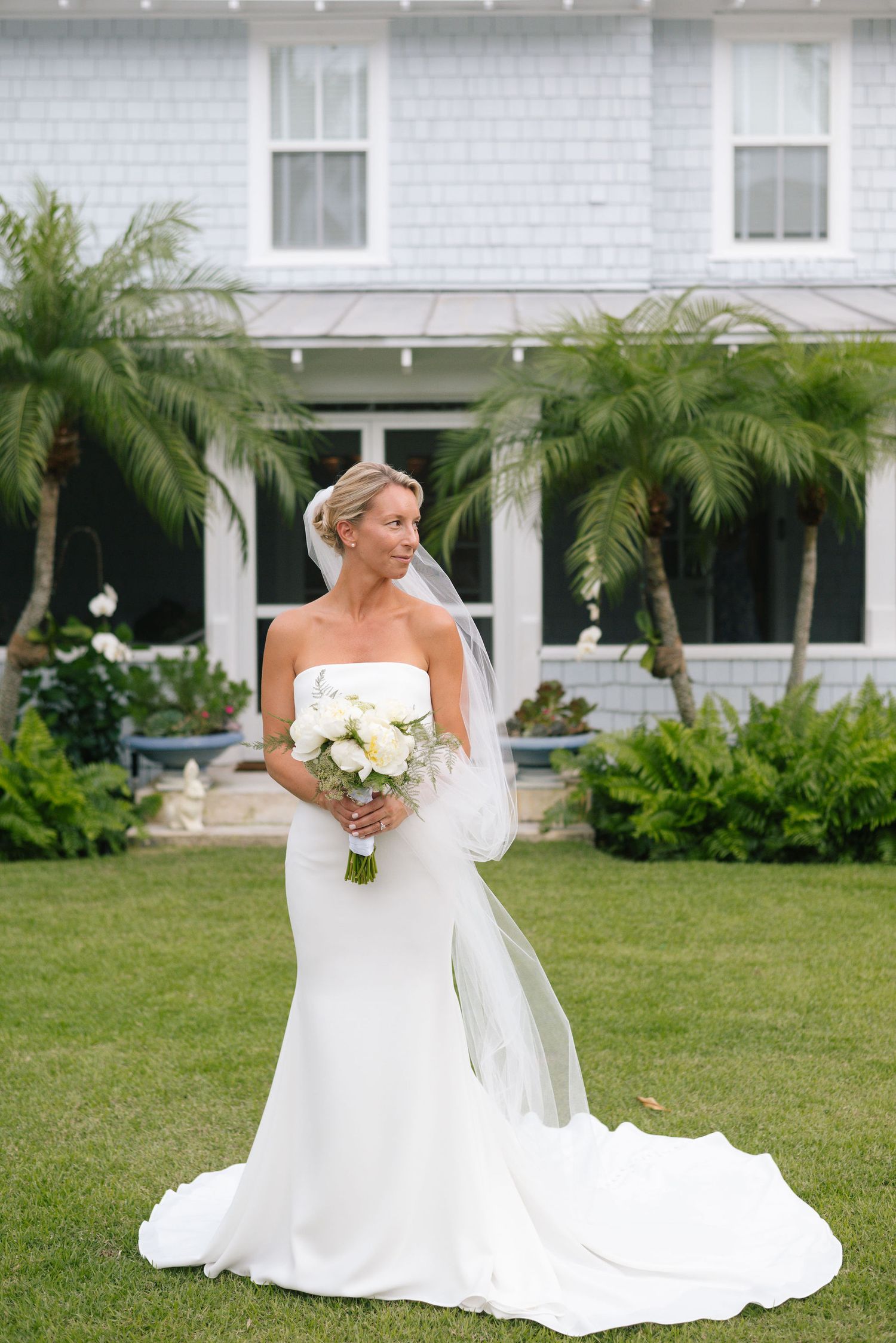 Bride in elegant white wedding gown poses gracefully on a lush green lawn with palm trees and white house in background.