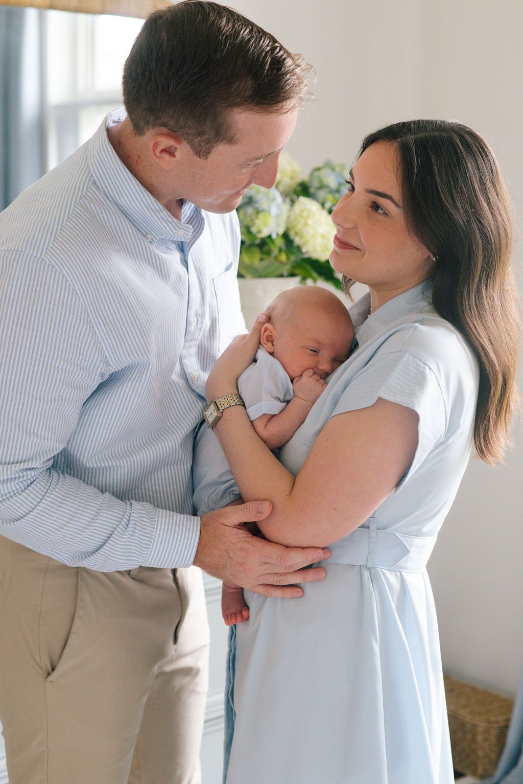 Parents share a loving moment while cradling their sleeping newborn baby in a sunlit room.
