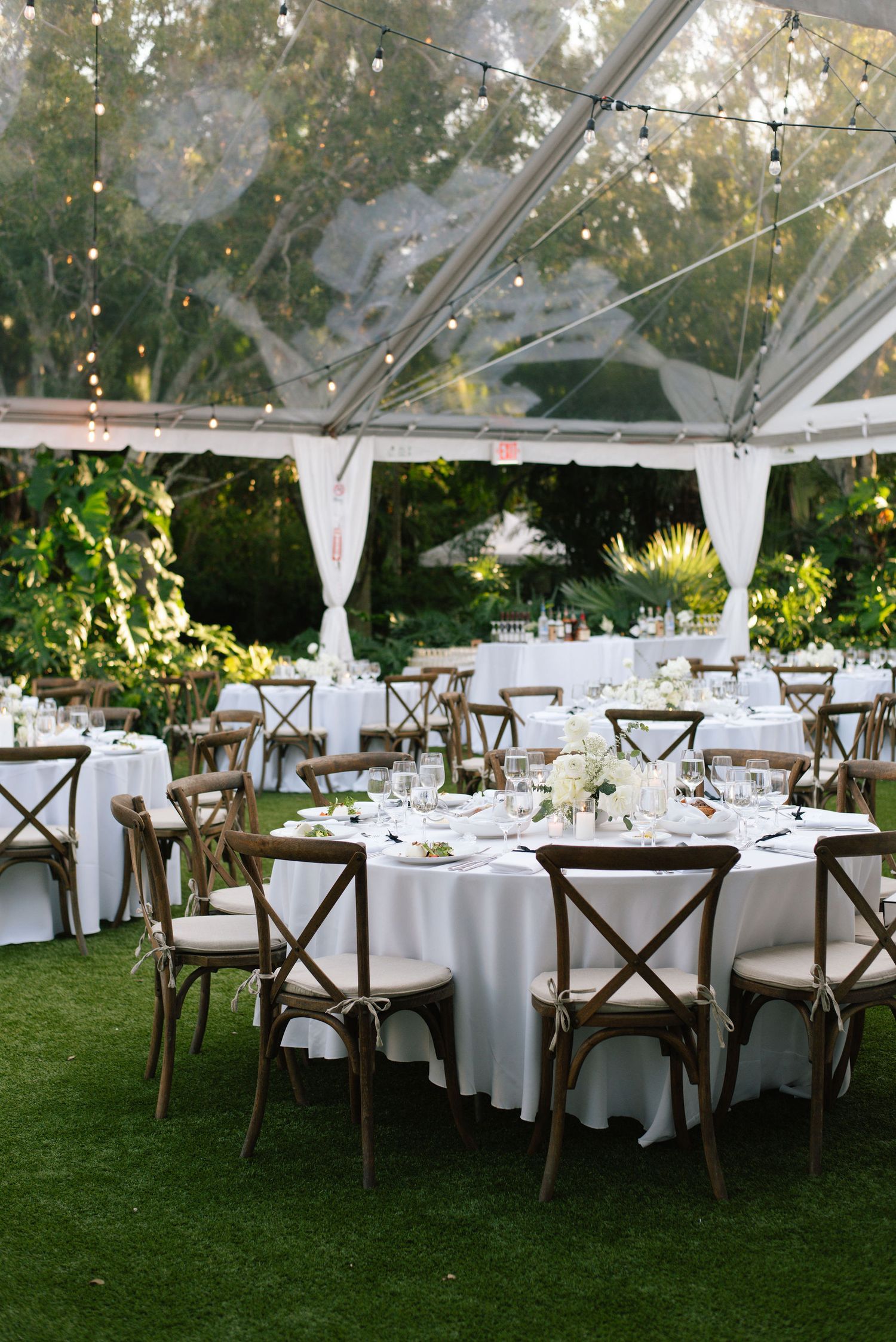 Elegant outdoor wedding reception setup with white tent, round tables, cross-back chairs and garden surroundings.
