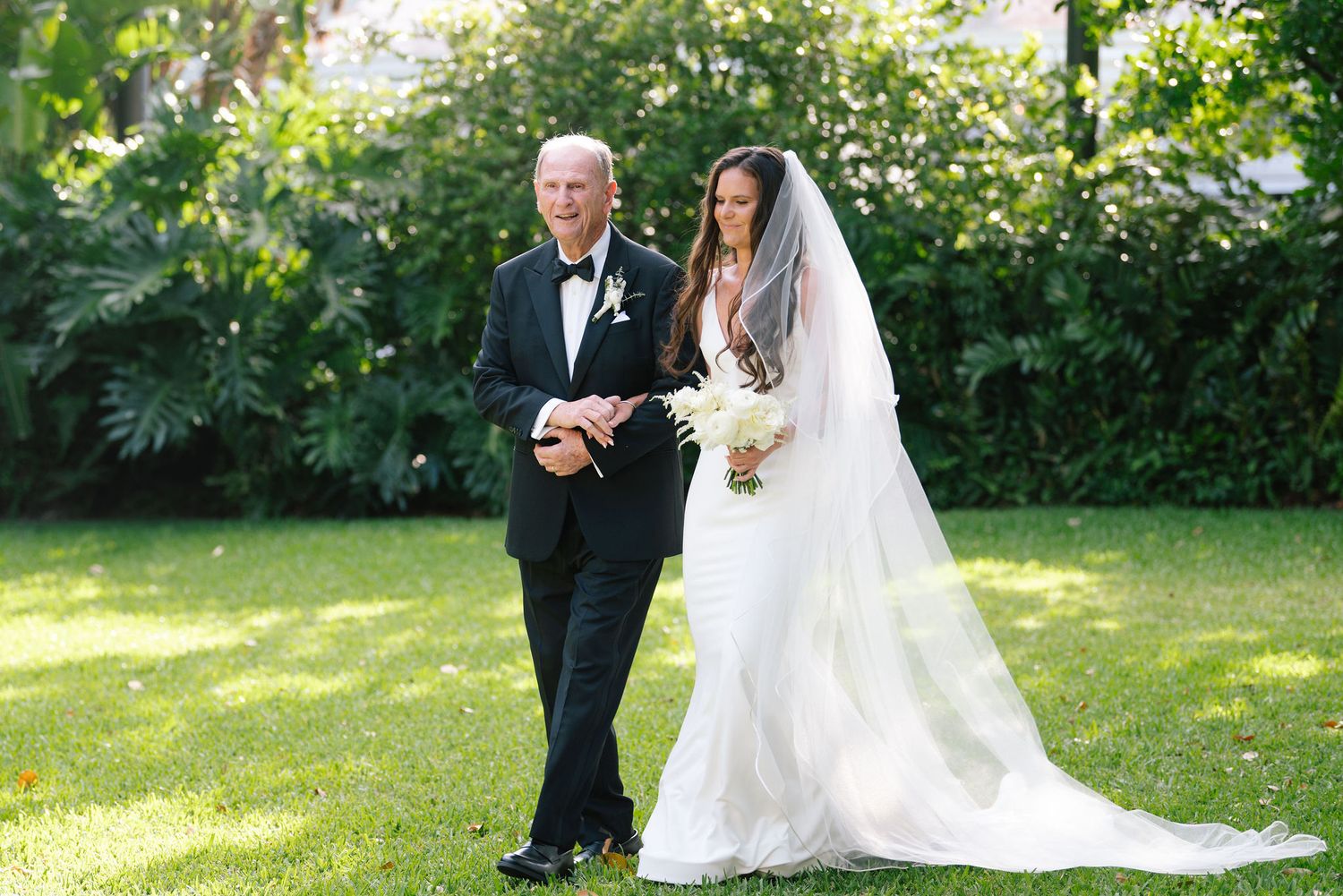An outdoor wedding ceremony takes place in a lush garden setting with white floral decorations.