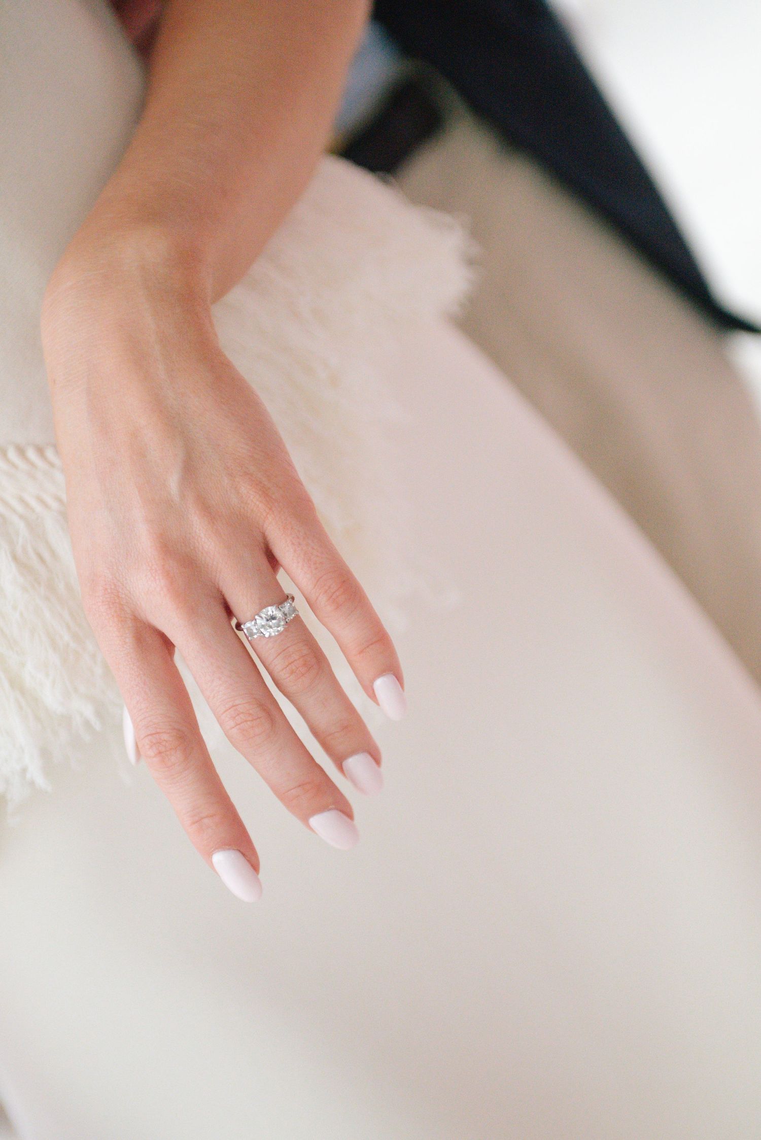 Delicate engagement ring sparkles on a manicured hand against white fabric.