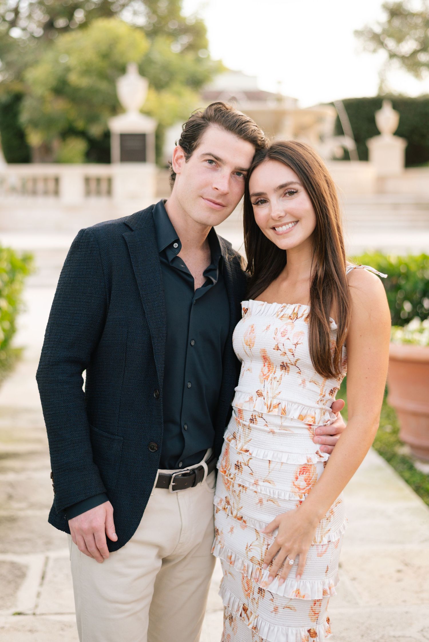 Couple poses together outdoors, with one wearing a dark blazer and the other in a white floral summer dress.
