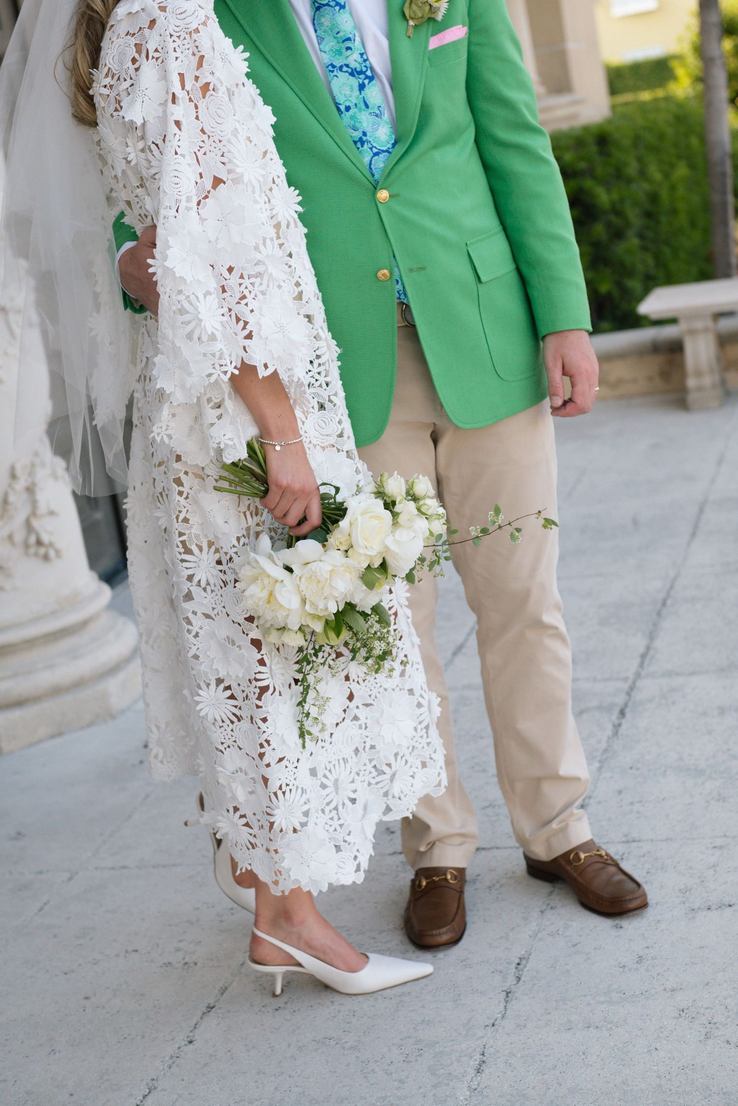Bride in white floral lace dress holding bouquet stands beside groom in green blazer and khaki pants outdoors.