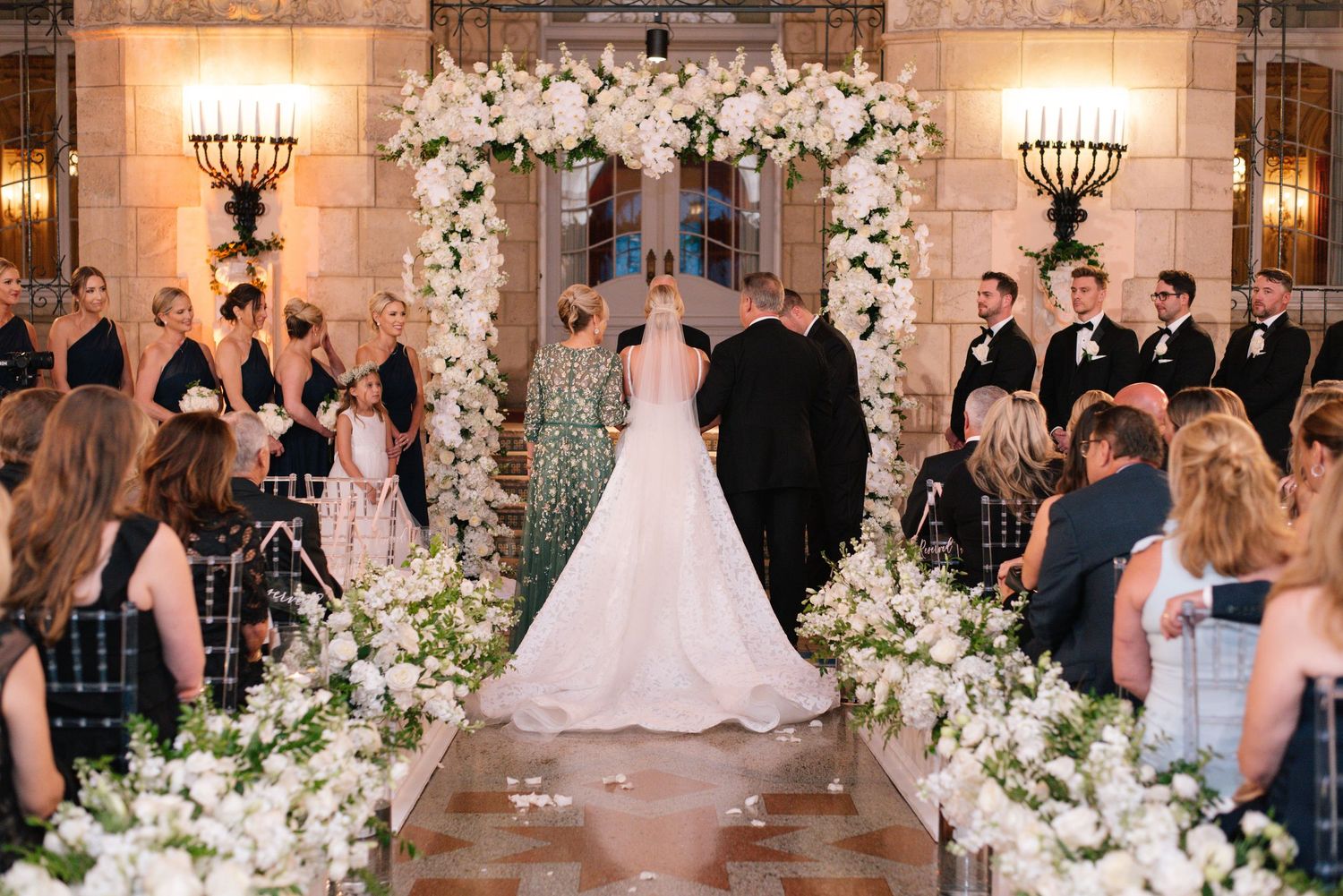 Bride walks down floral aisle toward white flower arch at elegant indoor wedding ceremony.