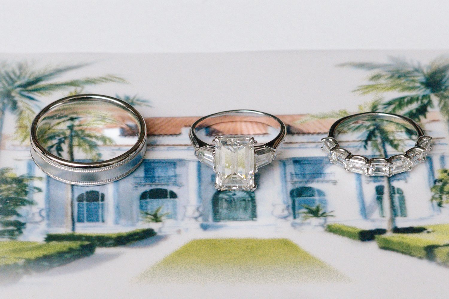 Three wedding rings displayed on a watercolor illustration of an elegant white mansion with palm trees.
