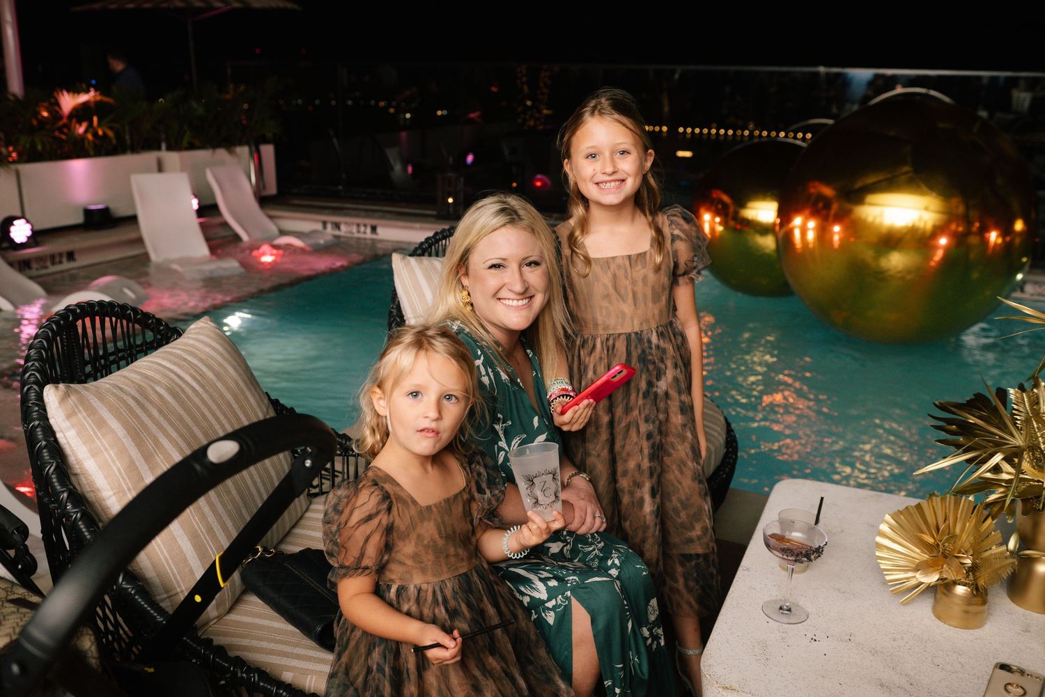 Festive nighttime party scene with people mingling around a swimming pool with glowing lights.