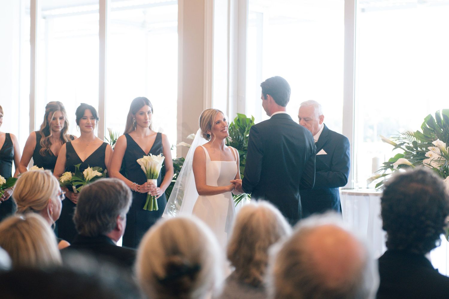Wedding ceremony guests seated in bright indoor venue with large windows.