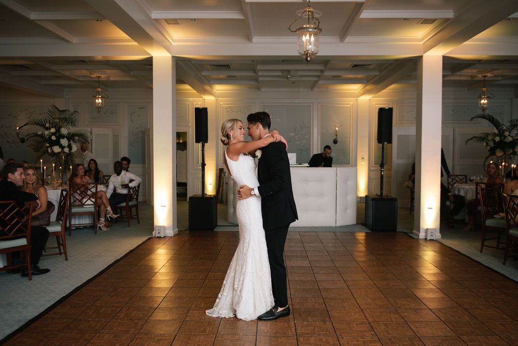 A couple shares their first dance in an elegant ballroom with white columns and warm lighting.