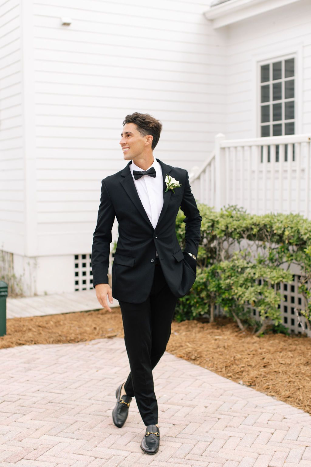 Groom in black tuxedo walking playfully on brick pathway.