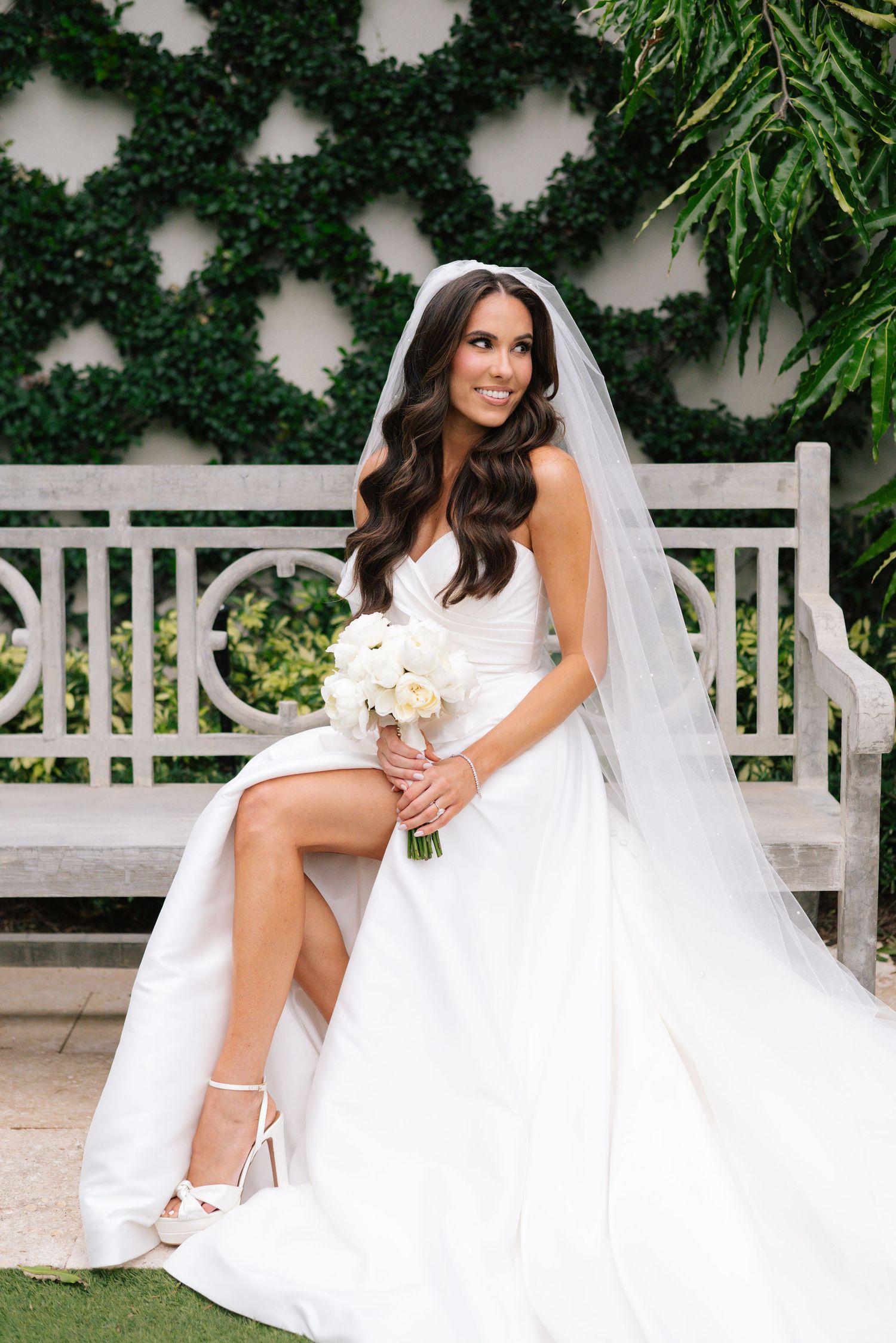 Bride in white dress poses on garden bench with lush greenery backdrop.
