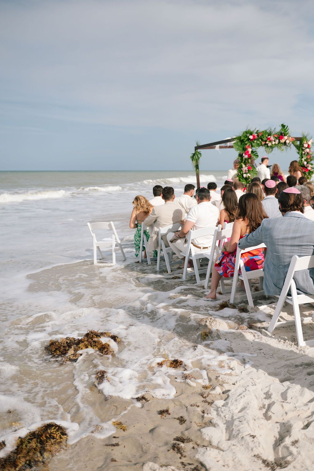 Waves crash near wedding ceremony setup with white chairs on sandy beach.
