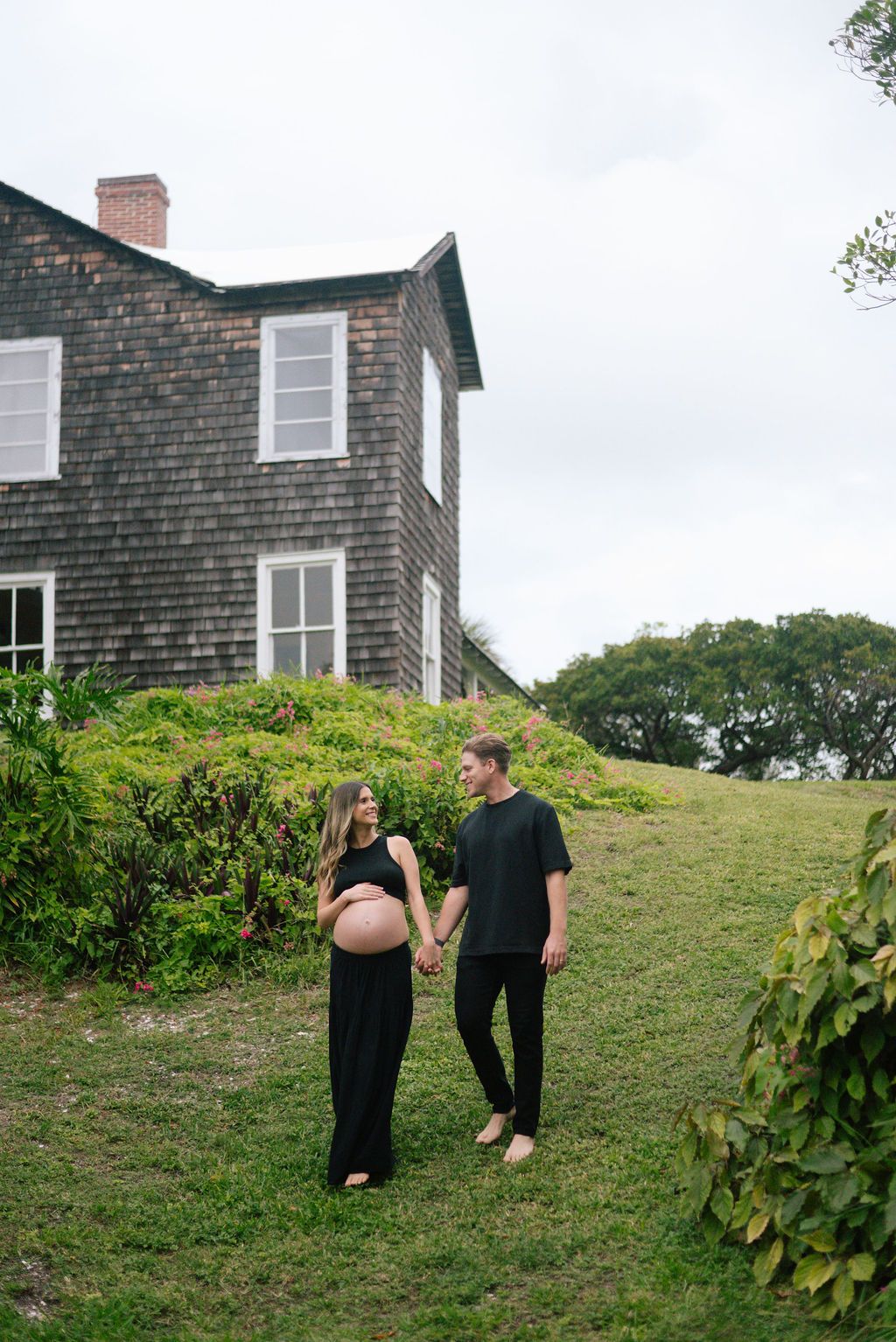 A couple in black outfits walks together on a grassy hillside near a shingled house.