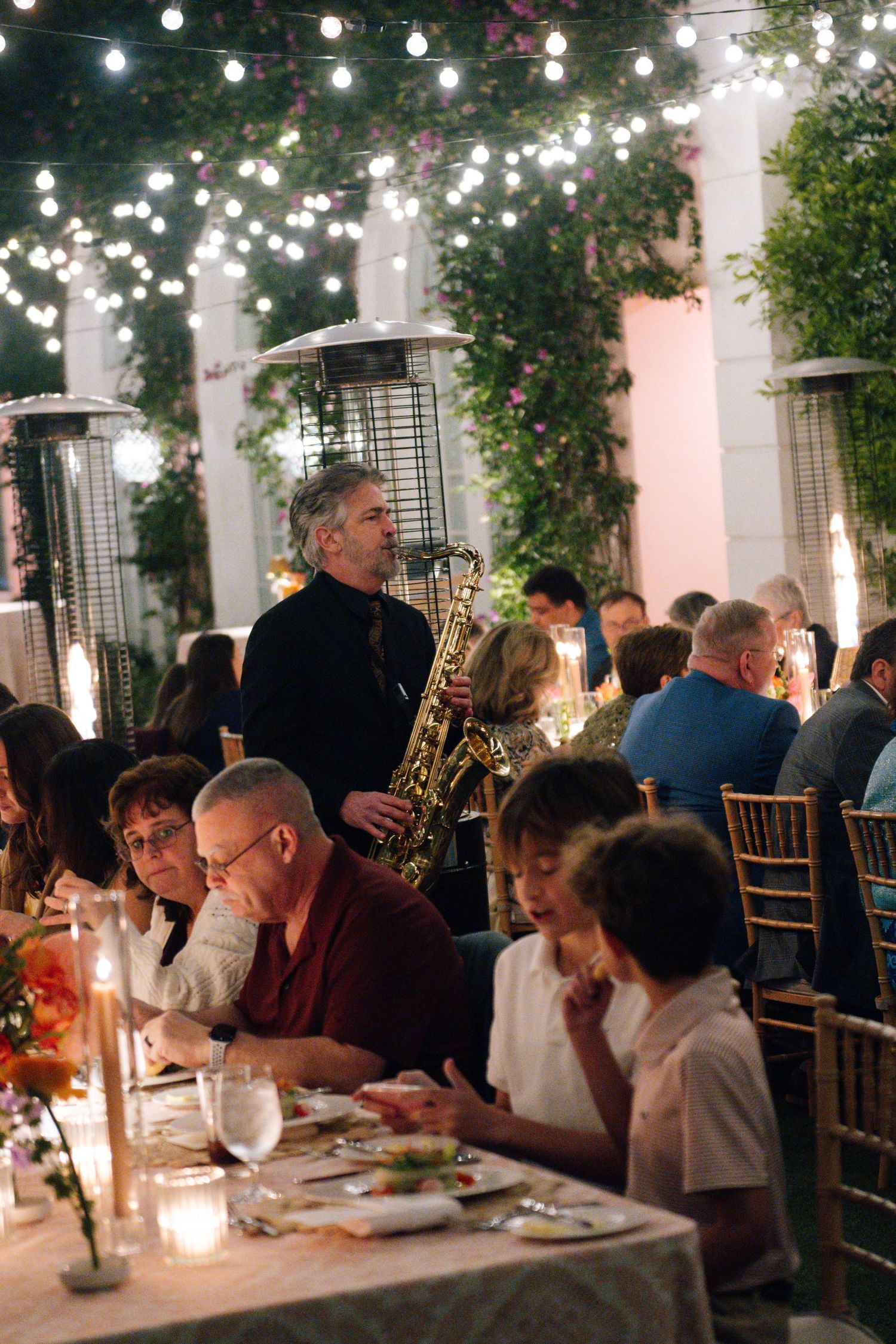 Guests enjoy an elegant outdoor dinner party under string lights with a saxophone musician performing.