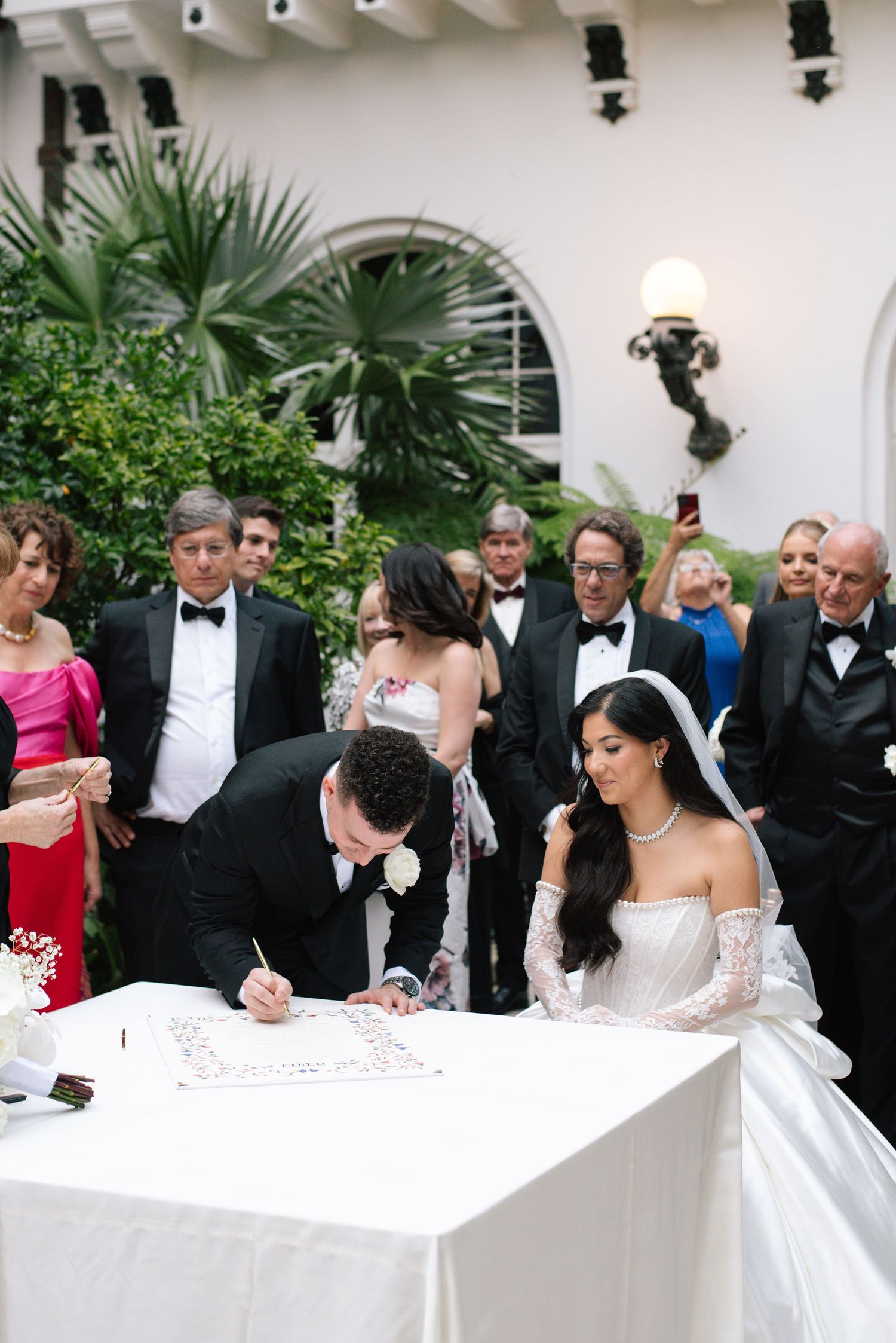 Couple signing marriage documents at a formal ceremony while guests look on at an elegant venue.