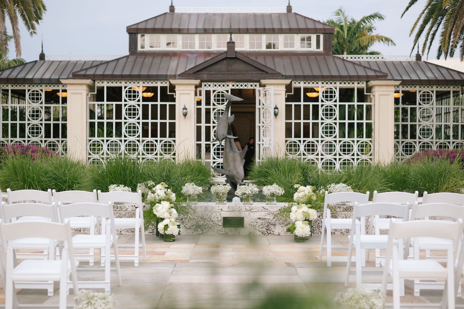White chairs arranged for an outdoor wedding ceremony with palm trees and a historic building in the background.