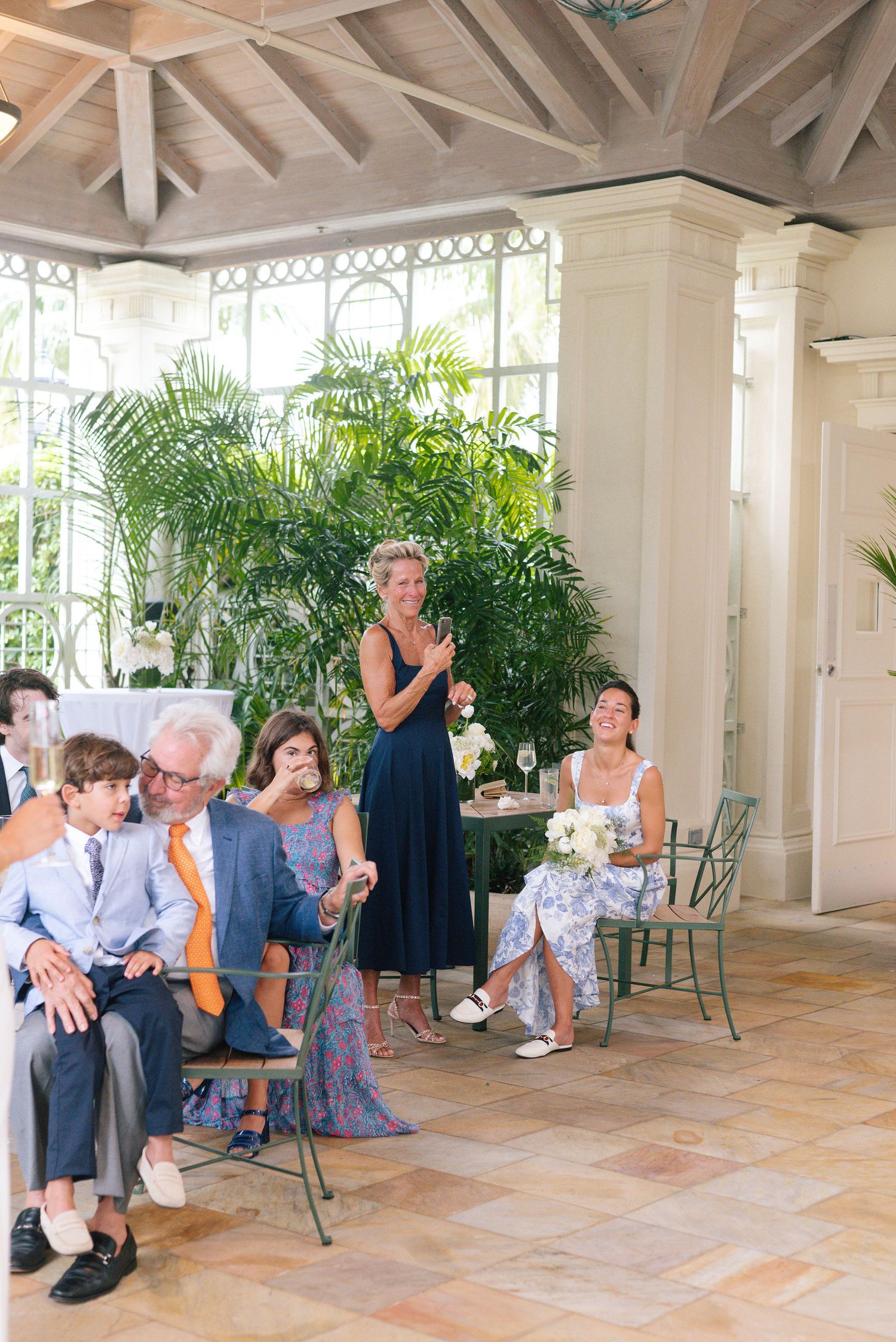 Wedding guests seated in a bright conservatory with tropical plants and cathedral ceiling.