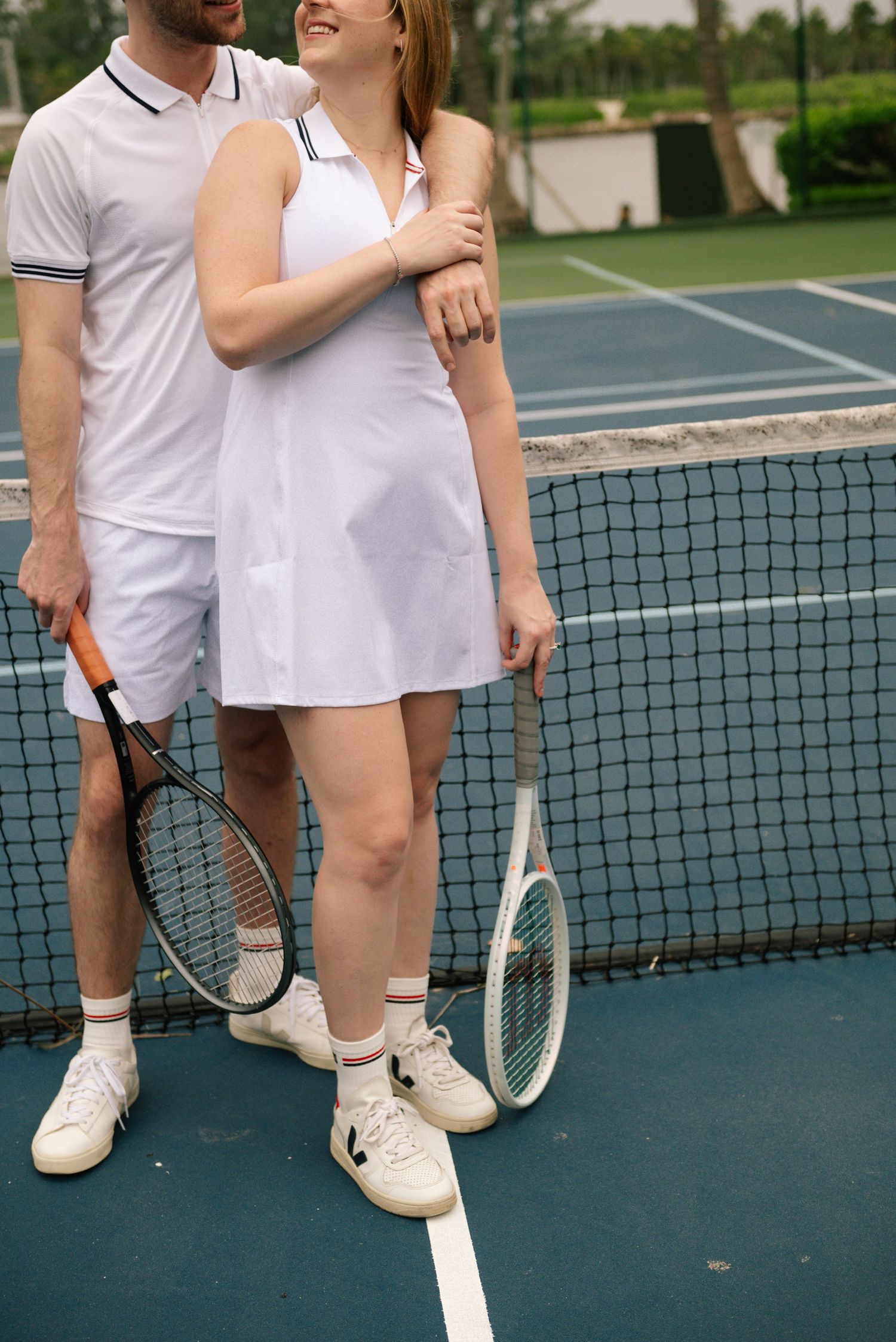 Tennis court engagement photos showing a couple in matching white outfits by the net.