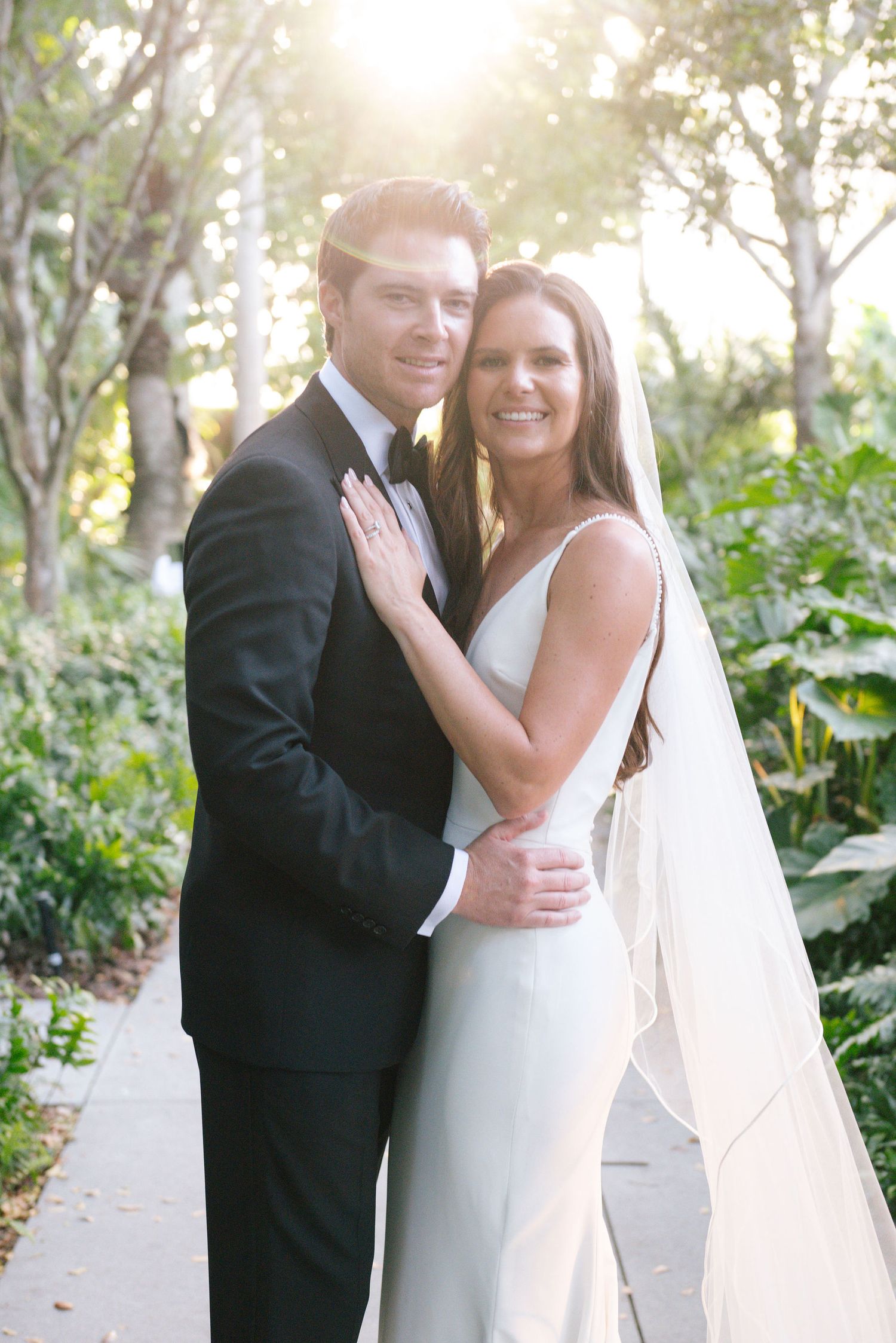 A bride and groom share romantic moments together in a lush garden setting with sunlit trees in the background.