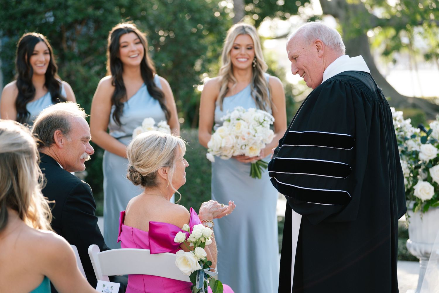 An outdoor wedding ceremony scene with guests and officiant gathered in a garden setting.