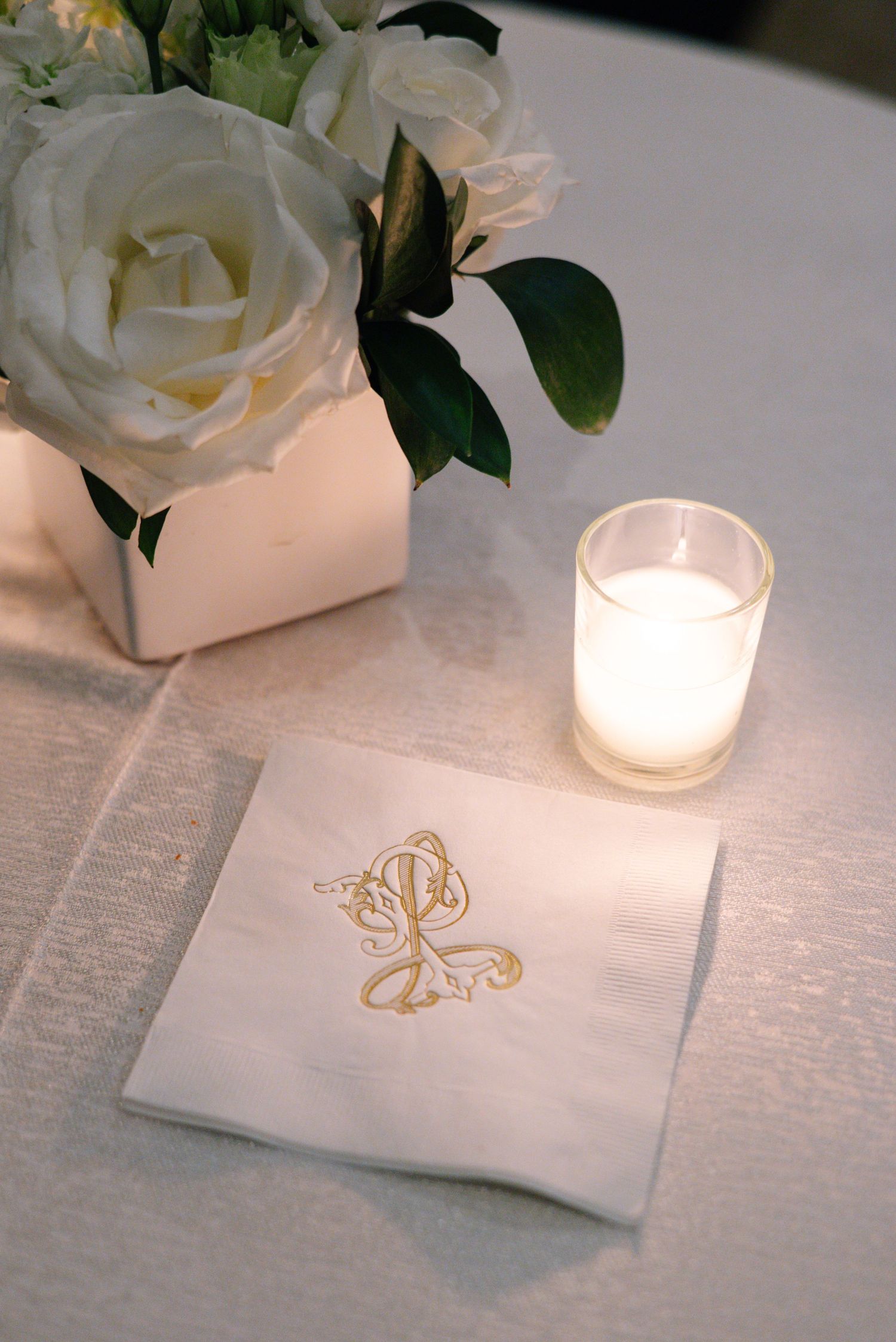 White rose centerpiece and monogrammed napkin with glowing votive candle on white tablecloth.