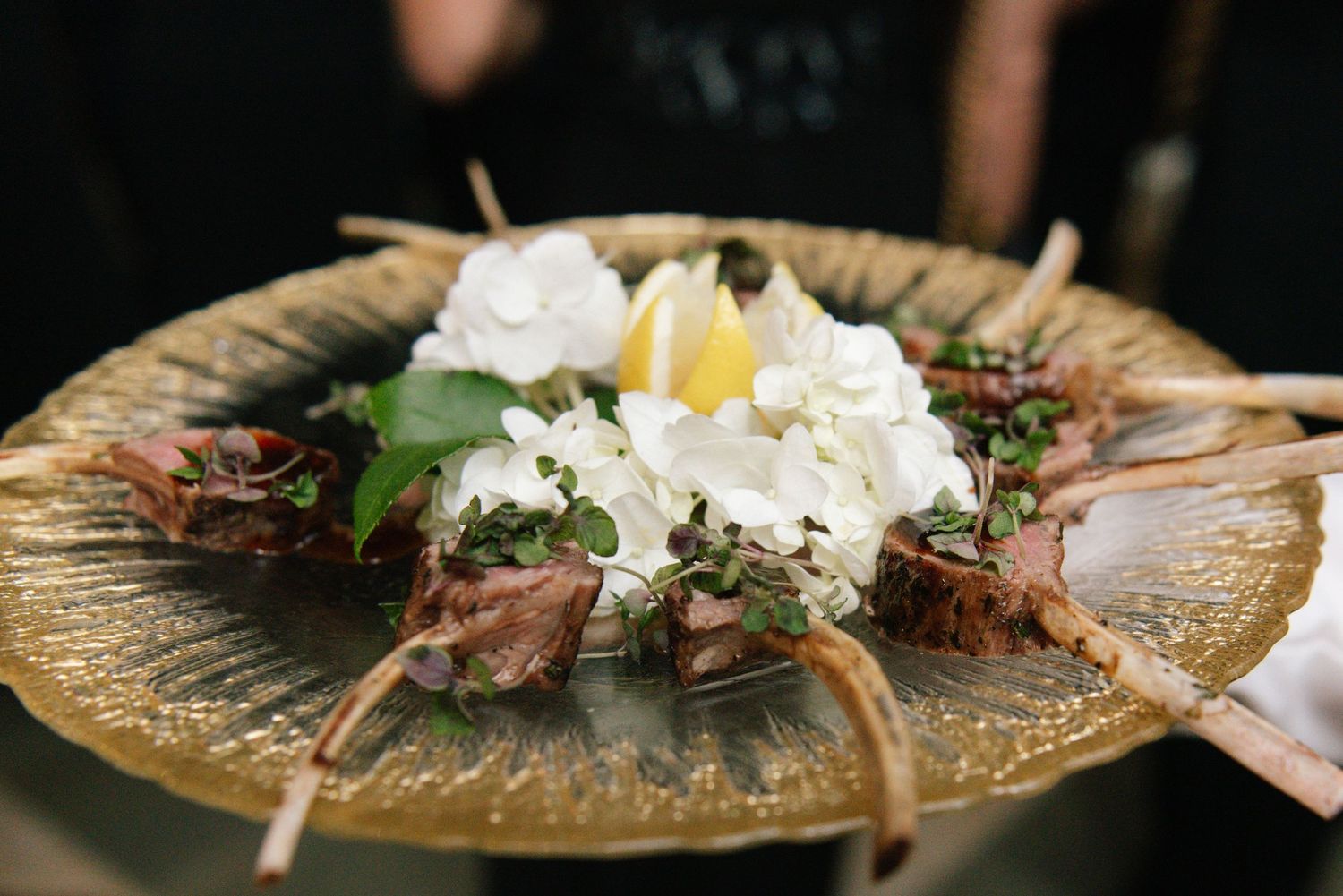 Elegant wedding appetizer platter featuring lamb chops arranged with white hydrangeas and lemon on a gold charger plate.
