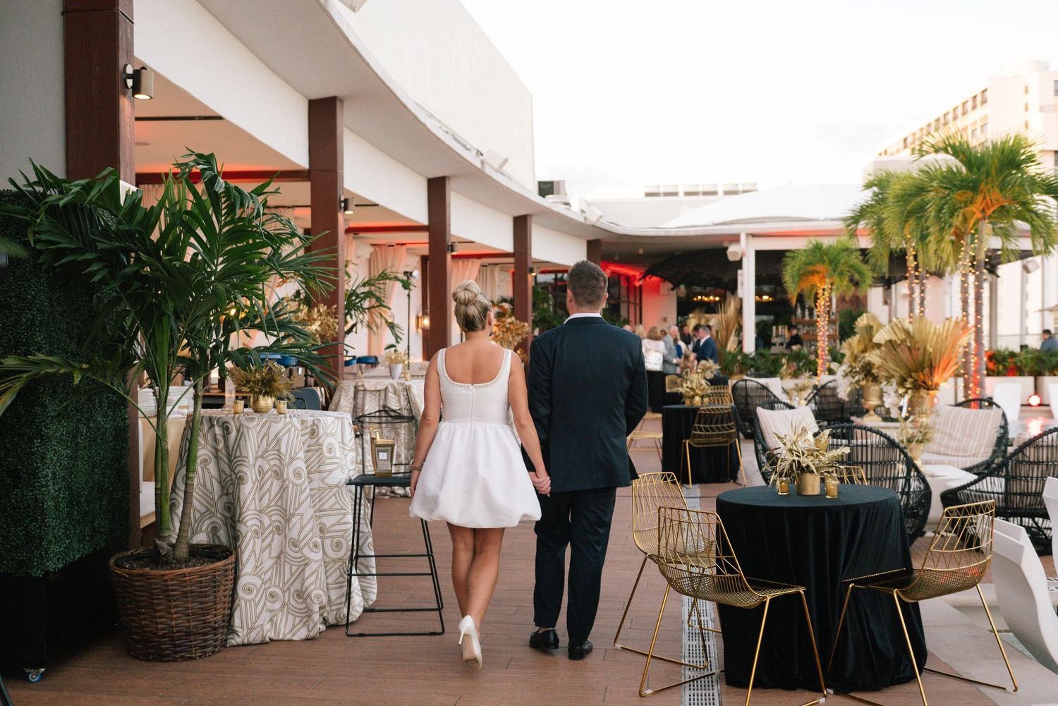 A couple walks along an elegant outdoor terrace decorated with palm trees and event furniture.