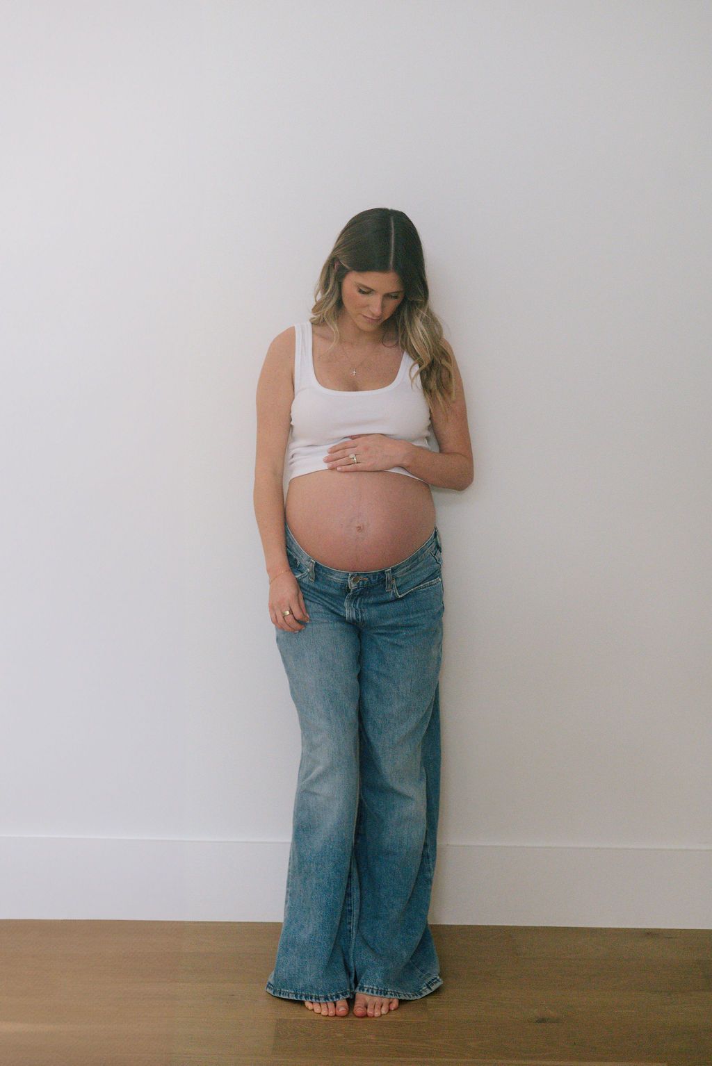 A maternity photo series showing a woman in casual attire against a white wall.