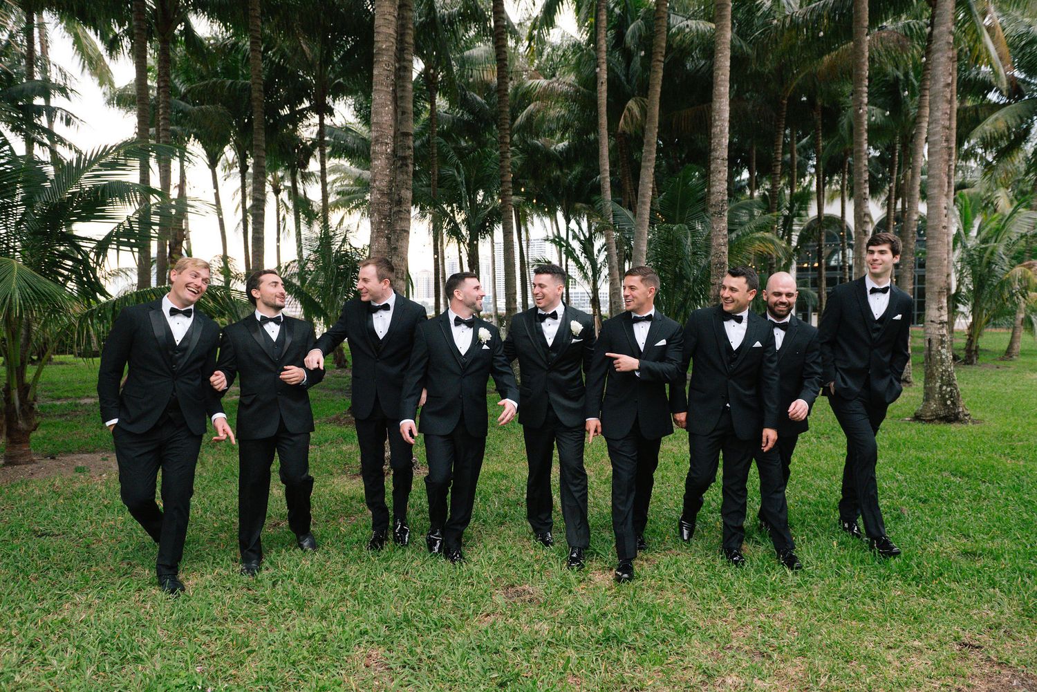 Wedding party in black tuxedos poses outdoors among palm trees at elegant venue.