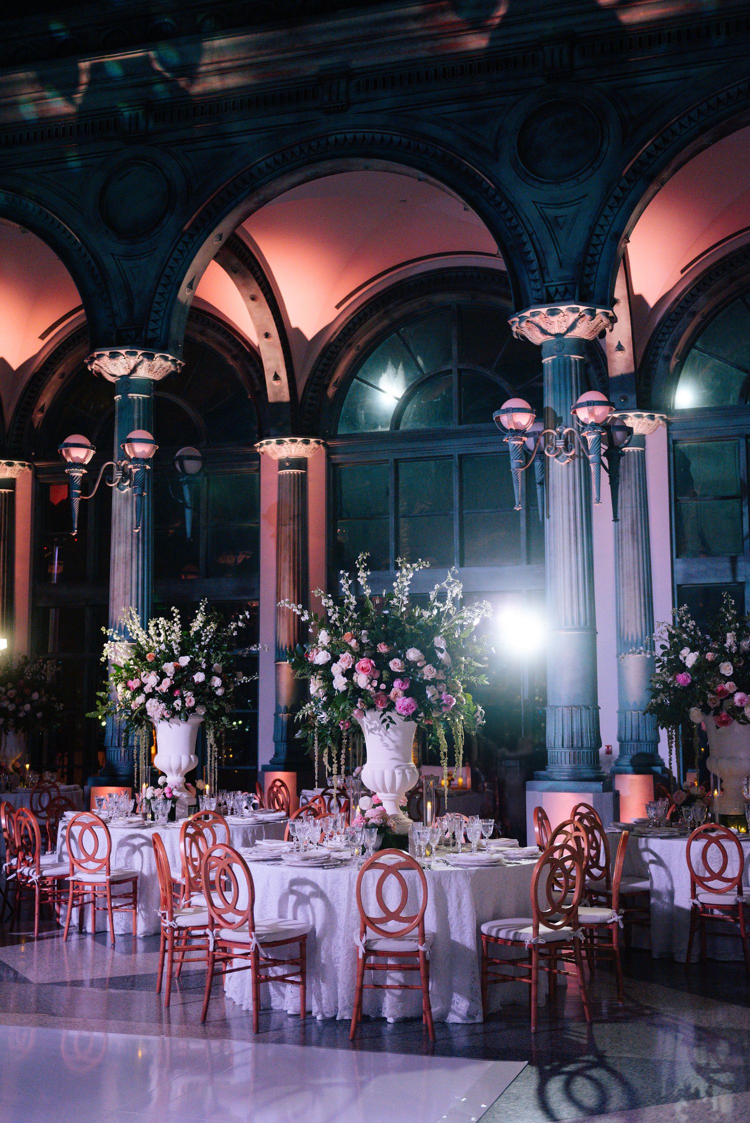 Opulent wedding reception venue with arched columns, floral arrangements, and pink ambient lighting.