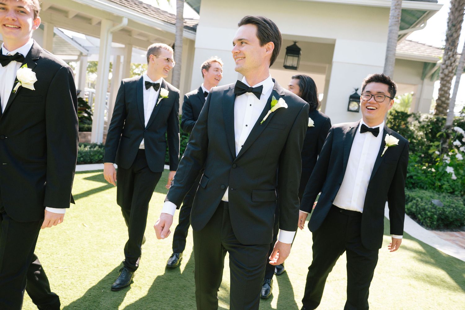 Wedding party members in formal attire stand together smiling outdoors.