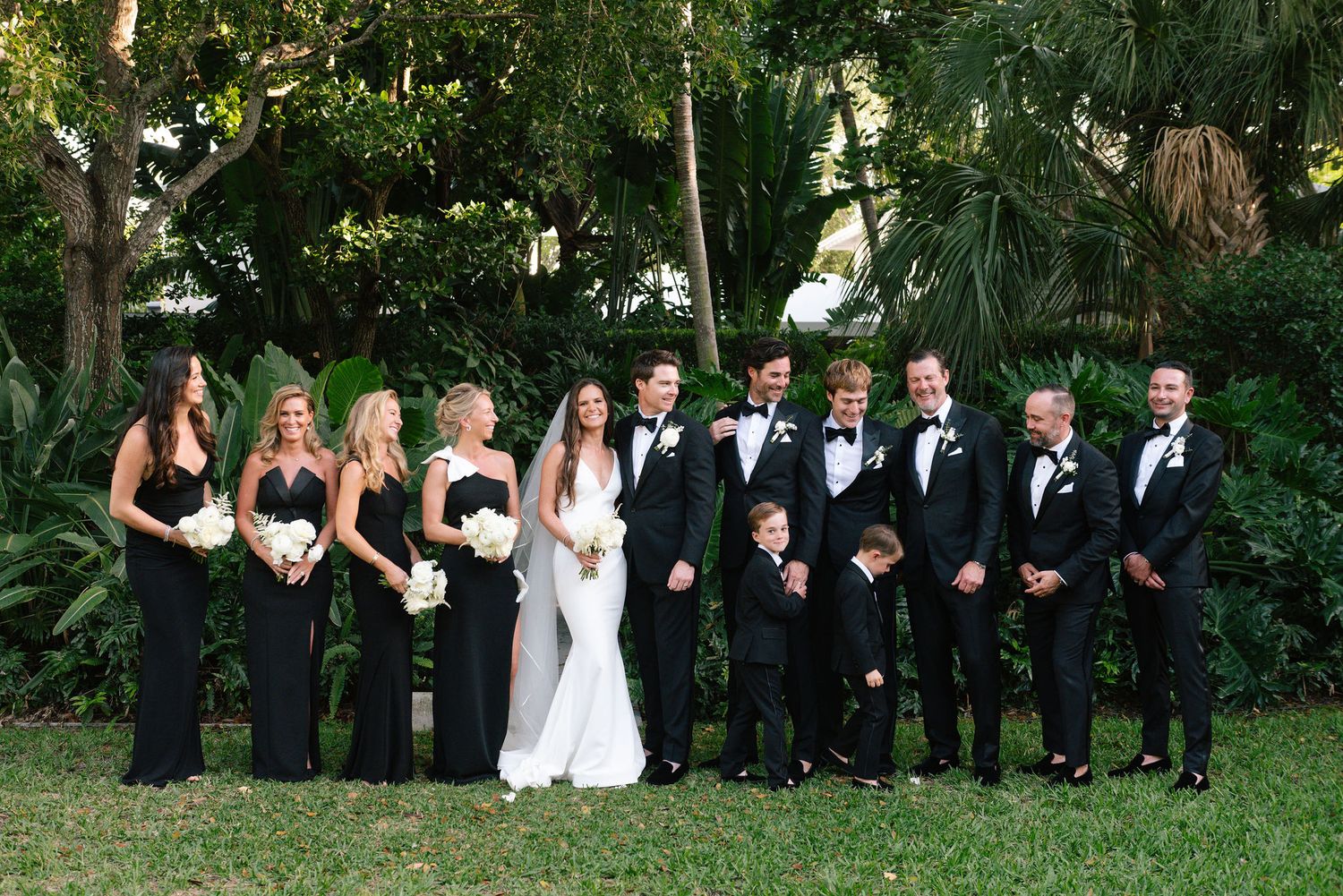 Wedding party in black formal attire poses together on green garden grounds.
