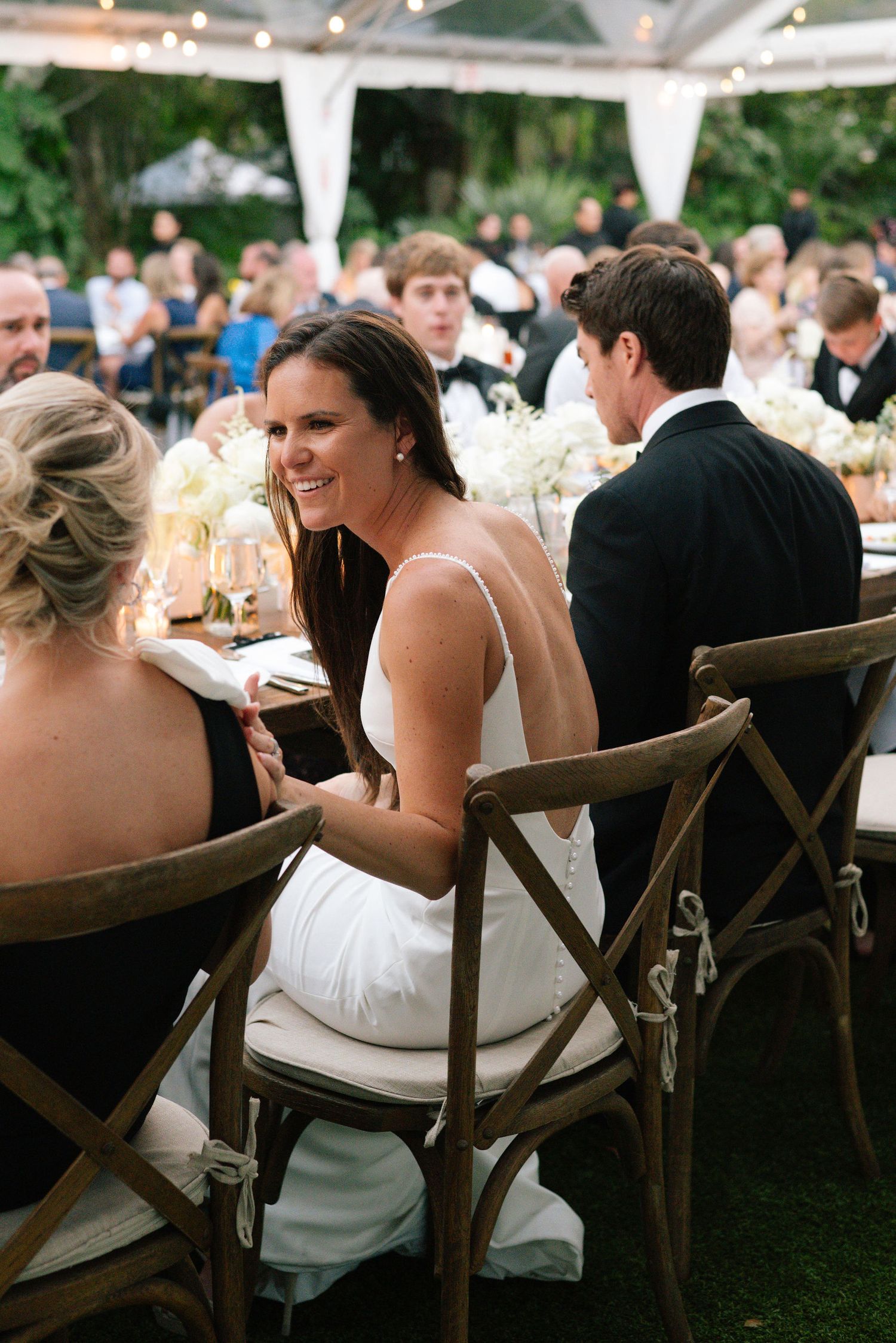 Outdoor wedding reception under a white tent with guests seated at wooden cross-back chairs.