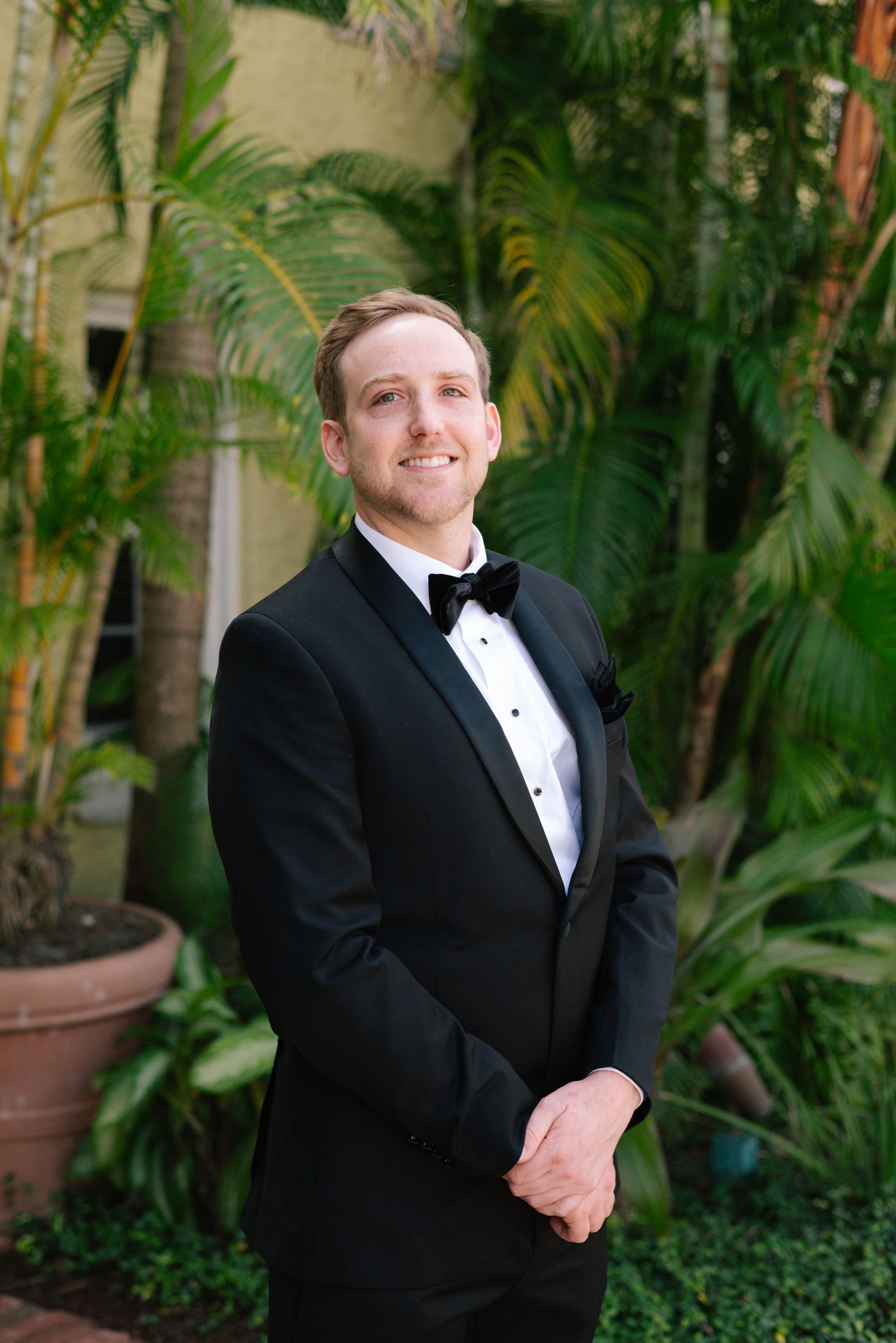 Man in formal black tuxedo smiles warmly against lush tropical garden backdrop.