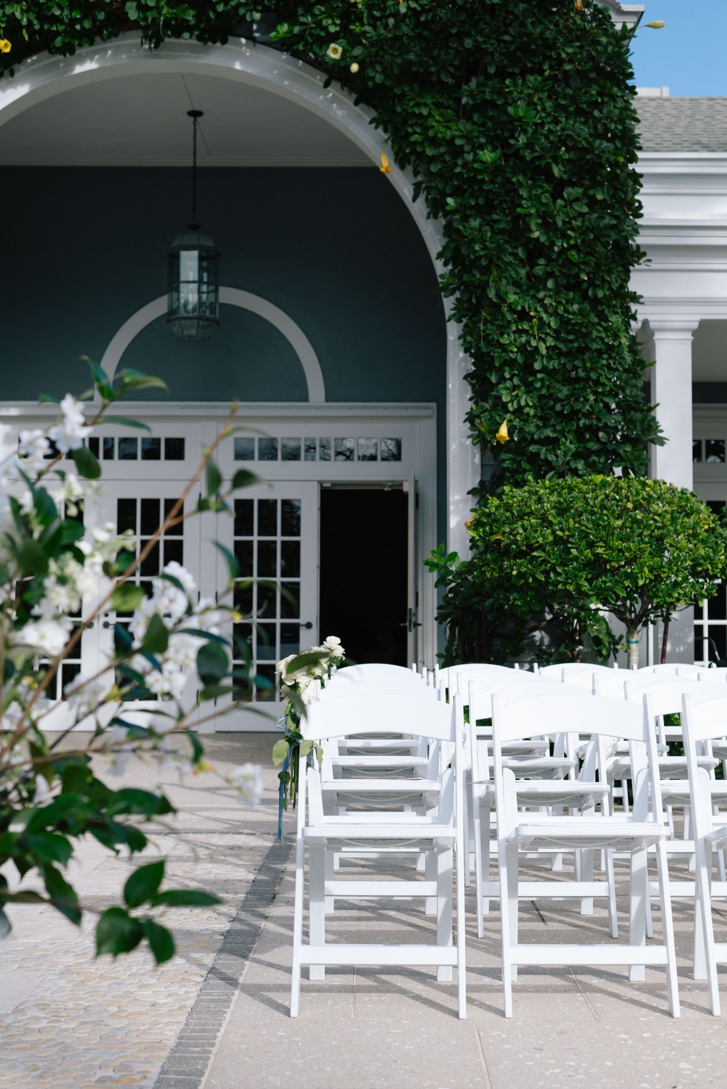 White wooden chairs set up for an outdoor wedding ceremony under elegant white archways with climbing vines.